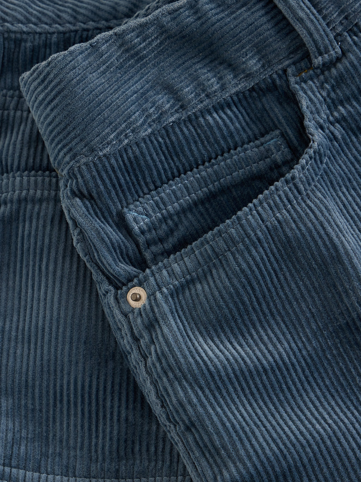 Close-up of a blue corduroy garment, showing the ribbed fabric texture and a small metal button near a pocket.