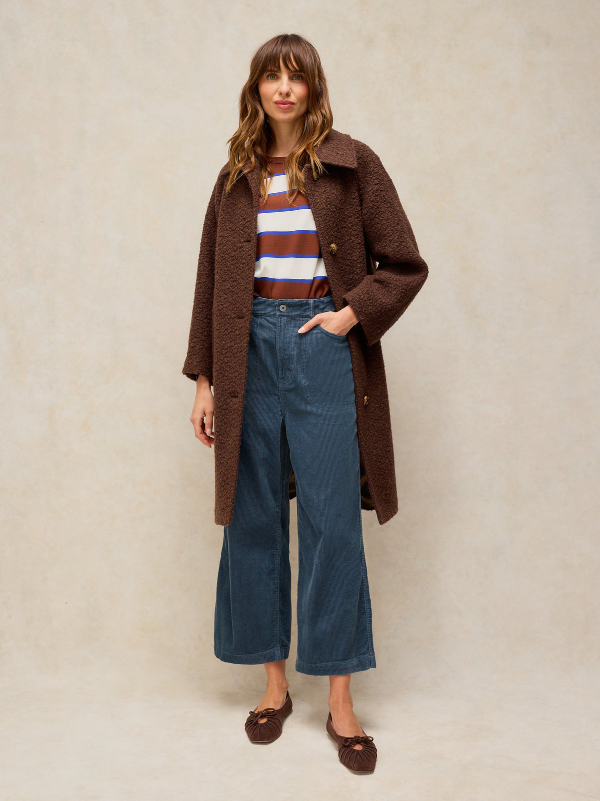 A woman stands against a textured beige studio backdrop wearing a brown teddy-textured coat over a striped top and blue wide-leg pants.