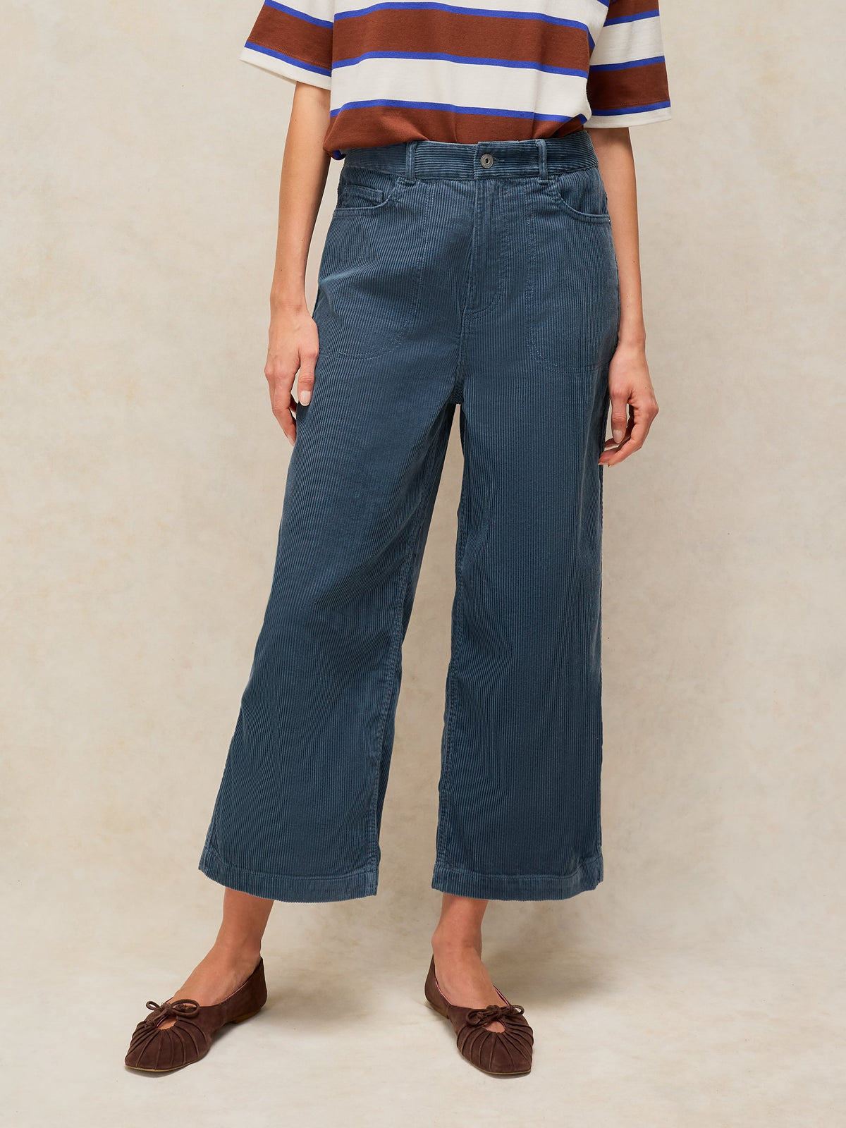 A model wearing a pair of dark blue cropped wide-leg jeans, paired with a brown, white, and blue striped t-shirt and brown flat shoes, standing against a textured beige studio backdrop.