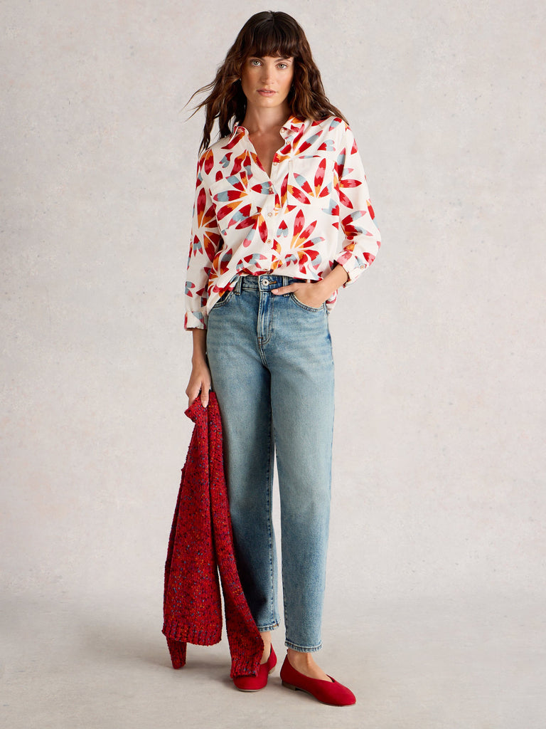 Woman wearing a colorful blouse, light blue jeans, and red shoes against a plain background