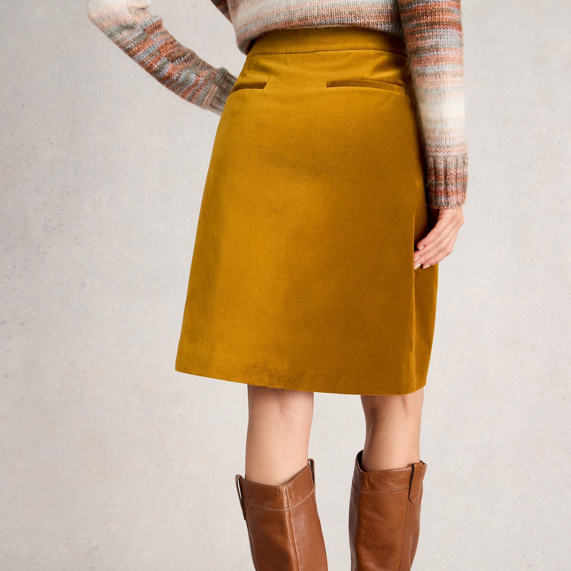Person wearing a mustard yellow skirt and brown knee-high boots on a plain background