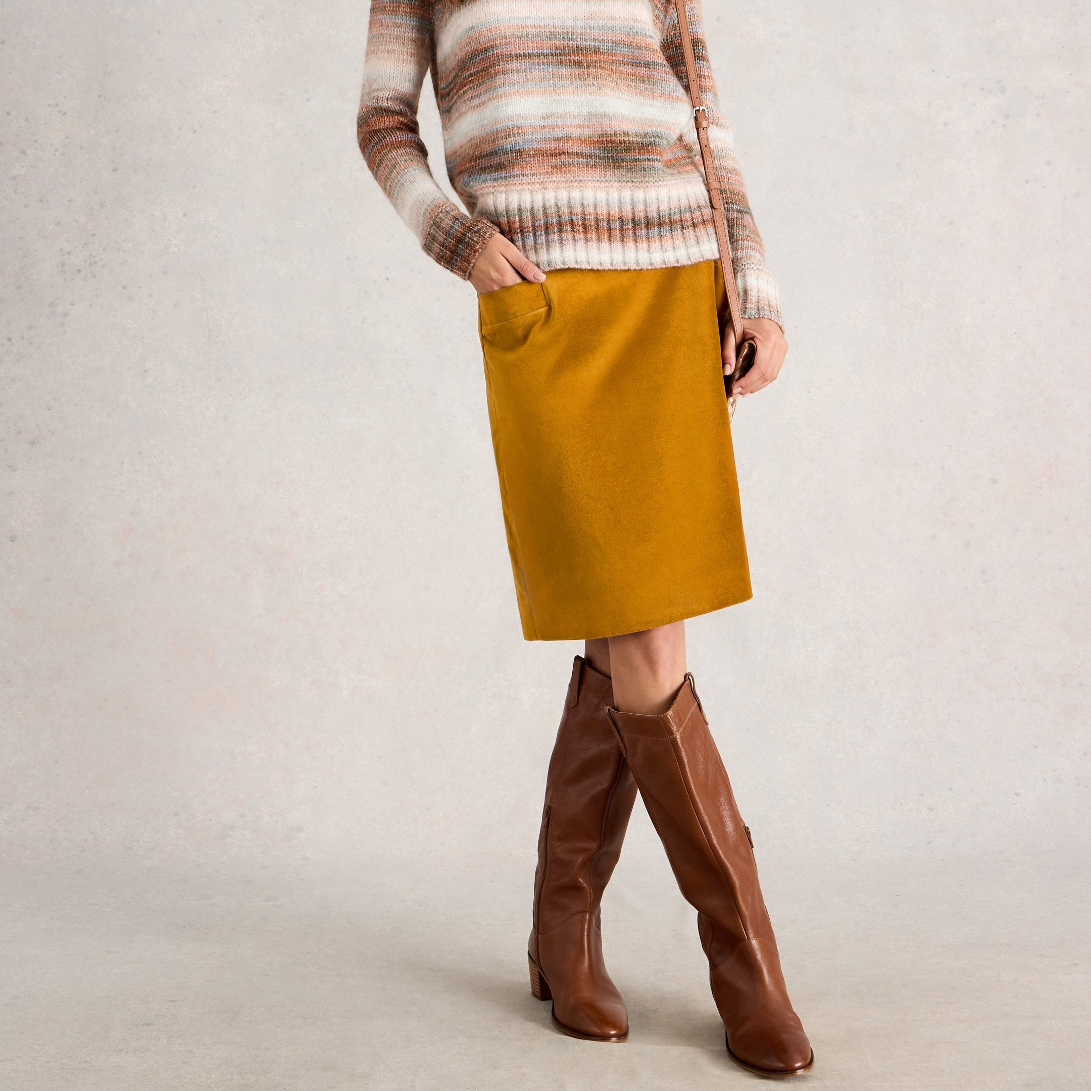 Woman wearing a striped sweater, mustard skirt, and brown knee-high boots on a plain background