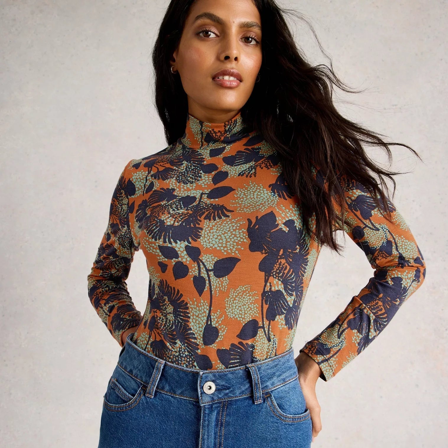 Woman wearing a patterned long-sleeve top and blue jeans against a plain background