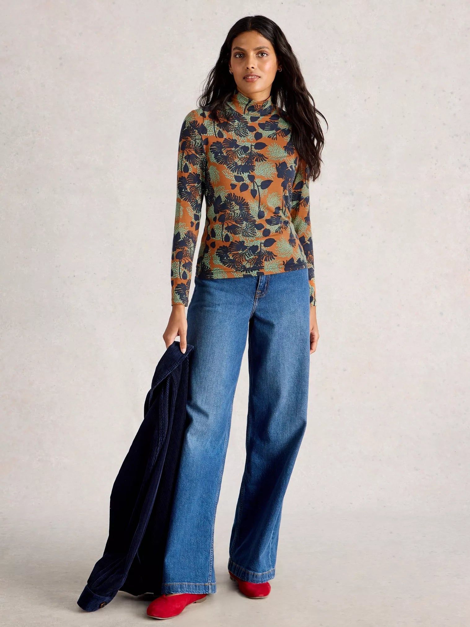 Woman wearing a patterned top and blue jeans holding a black jacket against a light background