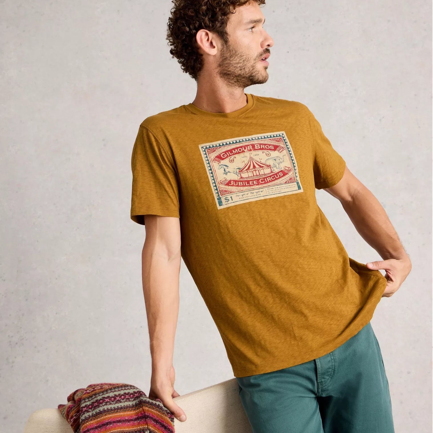Man wearing a mustard yellow t-shirt with a graphic design, standing next to a chair with a colorful blanket.