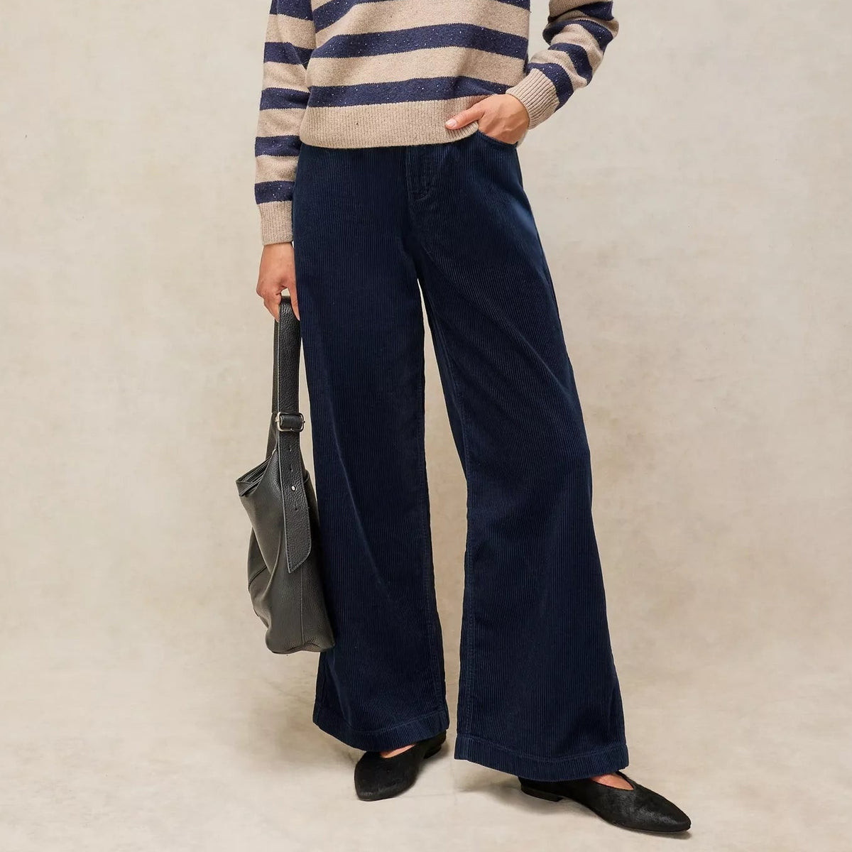 A woman stands against a warm beige studio backdrop wearing a beige and navy striped knit polo sweater, dark navy wide-leg trousers, black flats, and holding a gray shoulder bag.