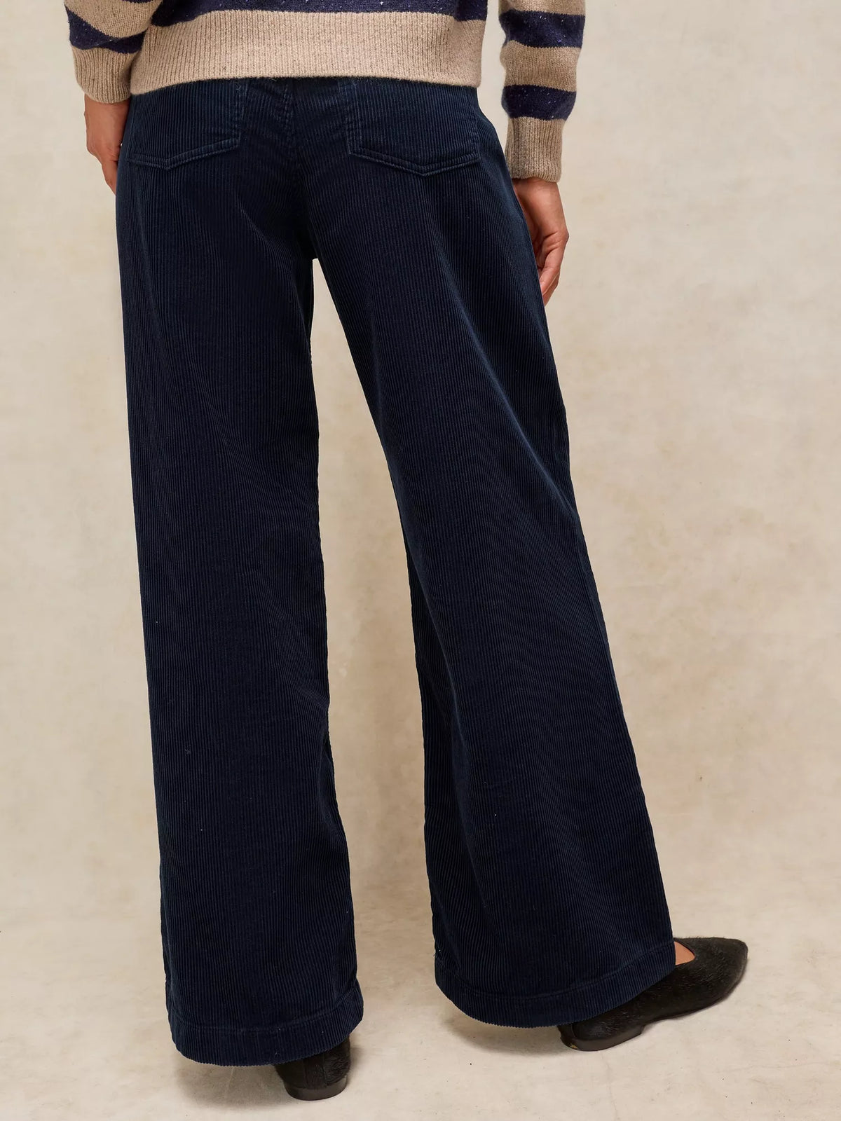 A person shown from the back wearing dark navy wide-leg corduroy pants, a beige and navy striped sweater, and black flats, standing against a textured beige studio backdrop.