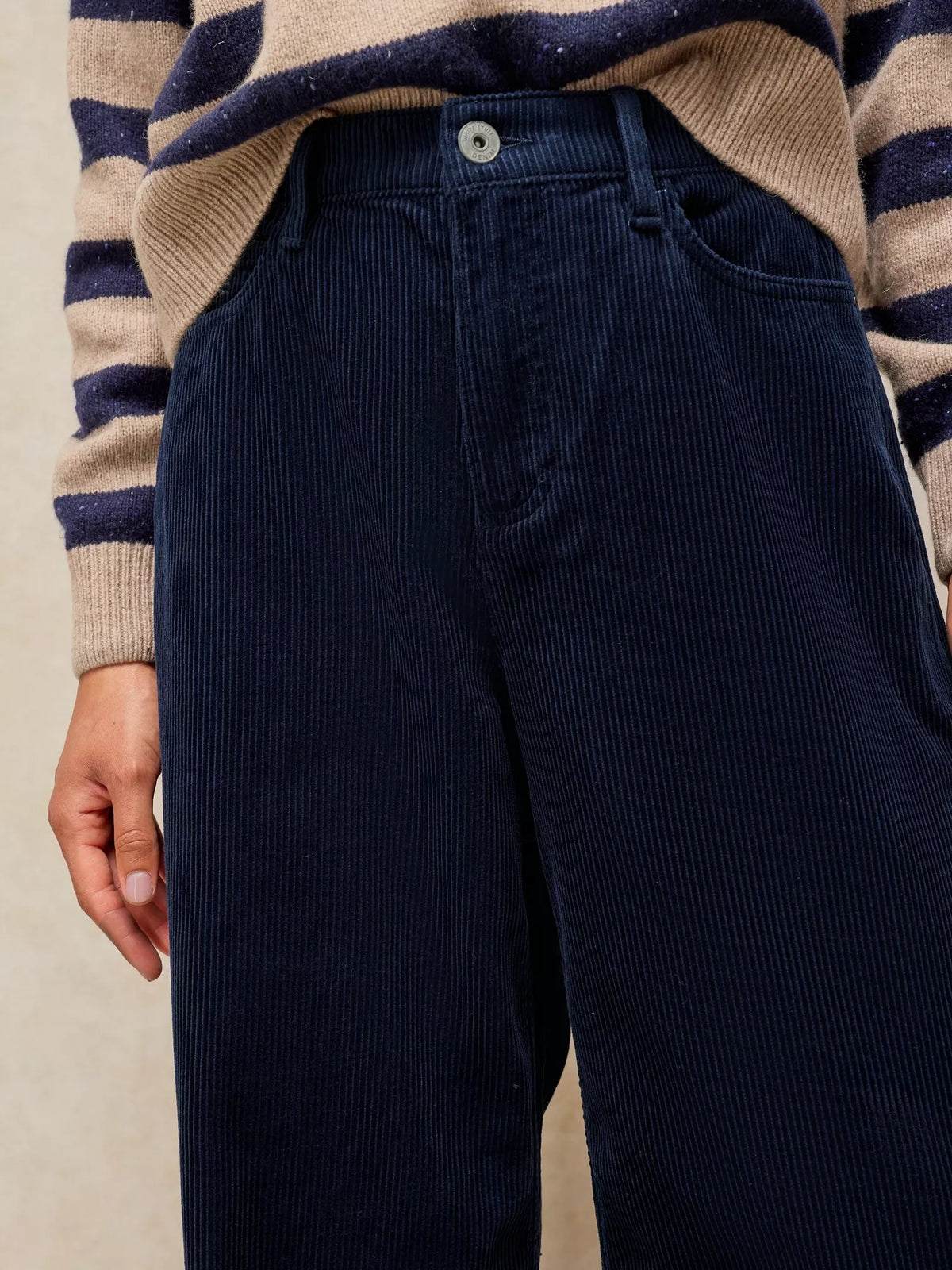 Close-up of a person wearing dark navy corduroy trousers and a beige and navy striped knit sweater, cropped from the chest down against a plain light beige background.