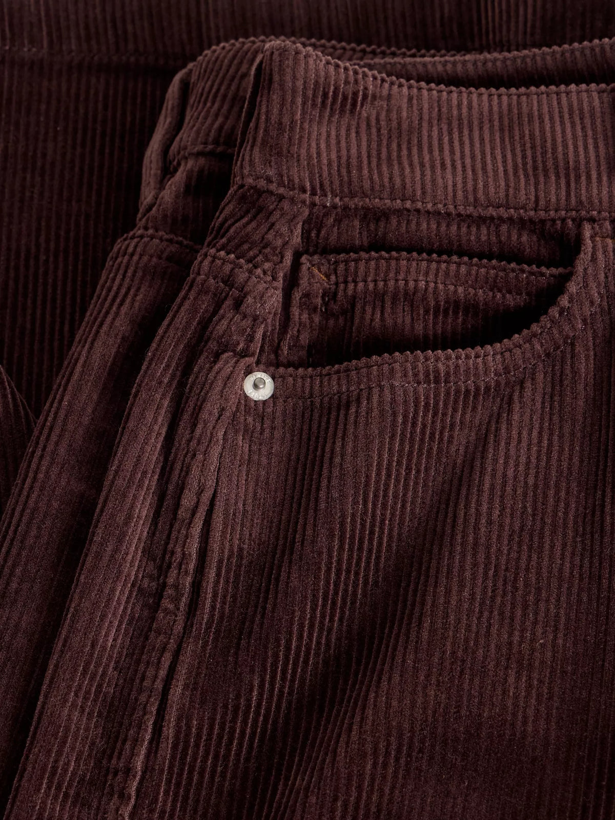 Close-up of dark brown corduroy trousers showing the ribbed texture, waistband, and a side pocket with a small silver button.