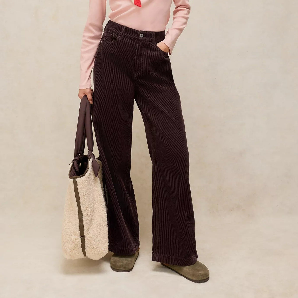 Woman wearing a pink blouse with a red scarf and dark pants, holding a beige bag on a plain background