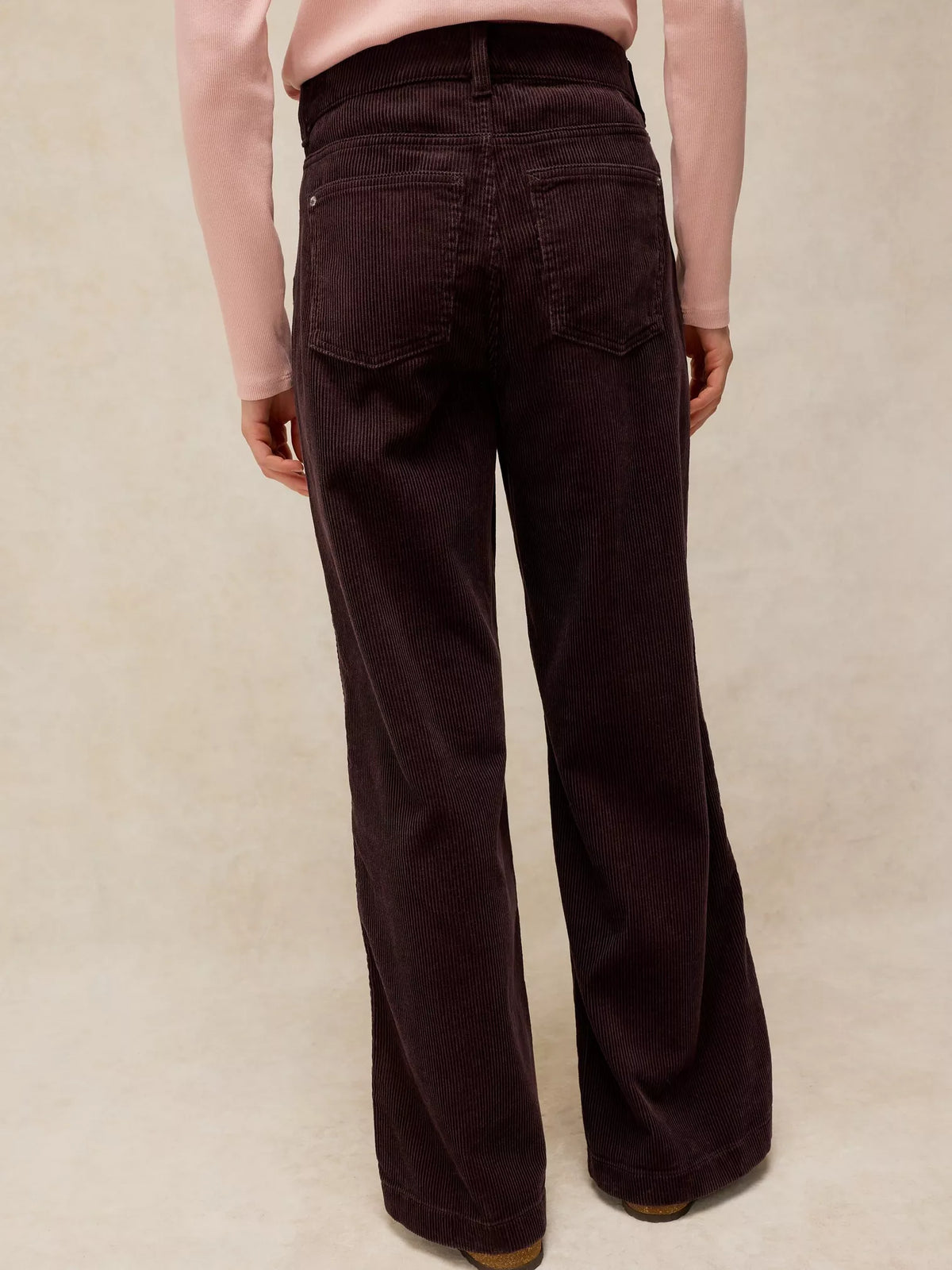 A person shown from the back wearing dark plum wide-leg corduroy pants and a light pink long-sleeve top, standing against a textured beige studio background.
