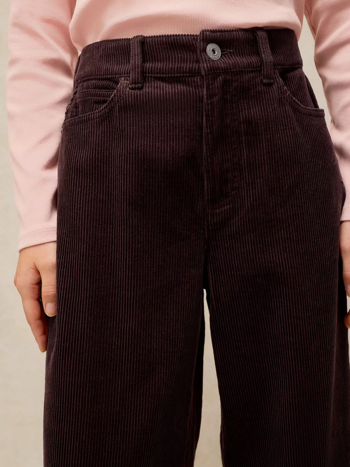 Close-up of a person wearing dark brown corduroy trousers with a pink long-sleeve top, showing the waistband, button, belt loops, and front pockets.
