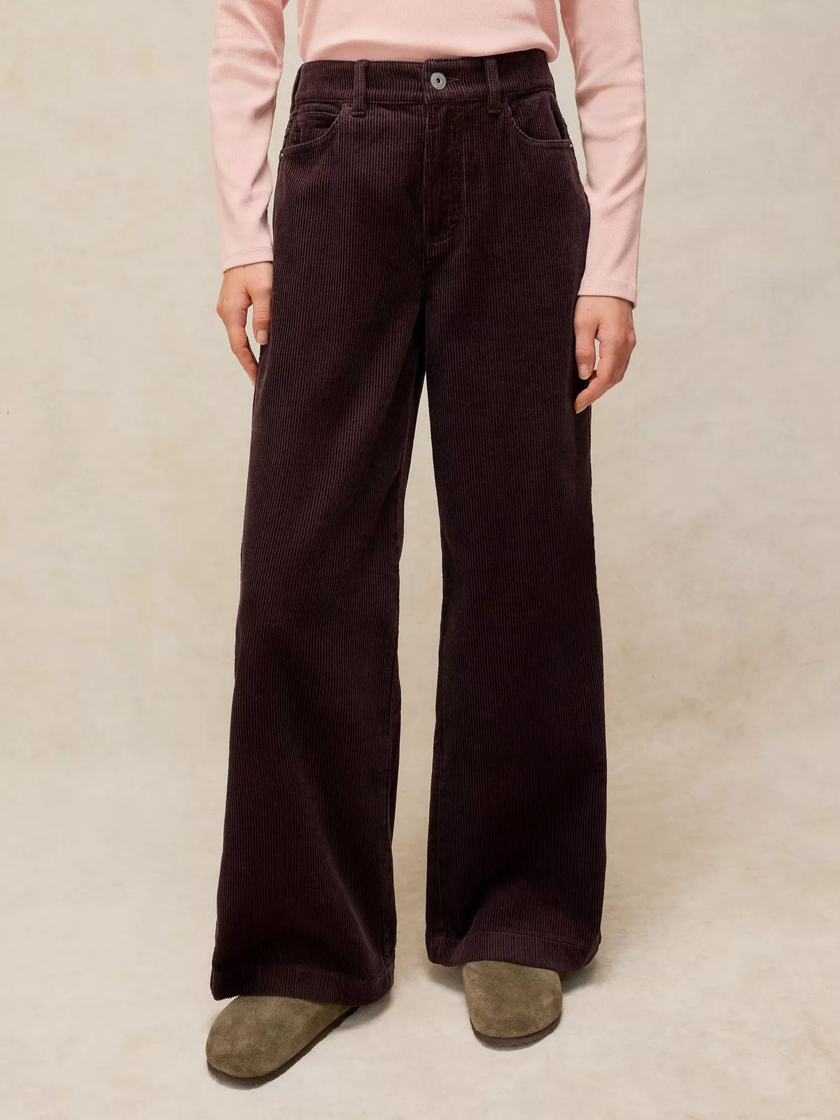 A person wearing dark brown wide-leg corduroy trousers, a light pink long-sleeve top, and brown suede shoes, standing against a neutral beige textured background.