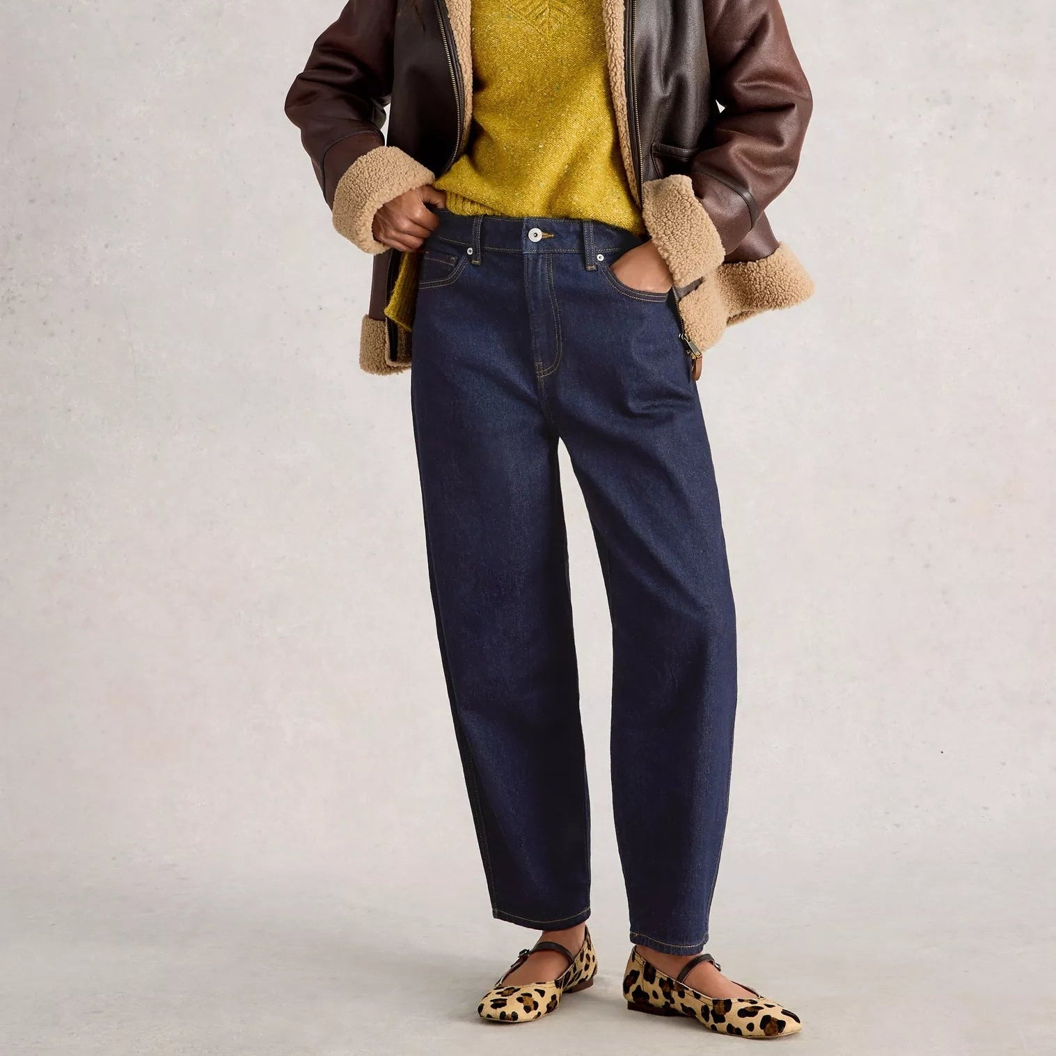 Woman wearing a brown shearling jacket, yellow sweater, and blue jeans on a light gray background