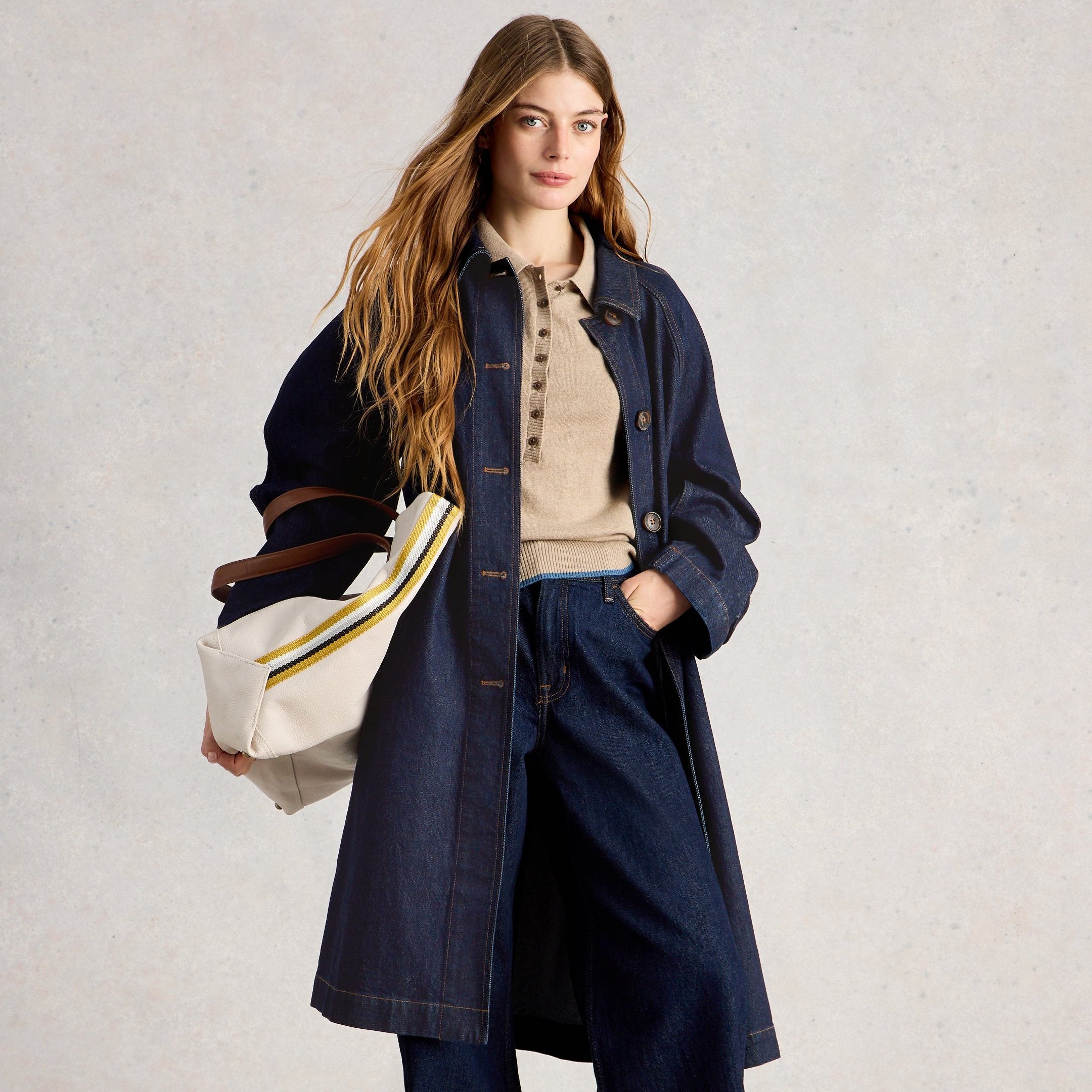 Woman wearing a long navy coat holding a white bag with a striped handle on a light background