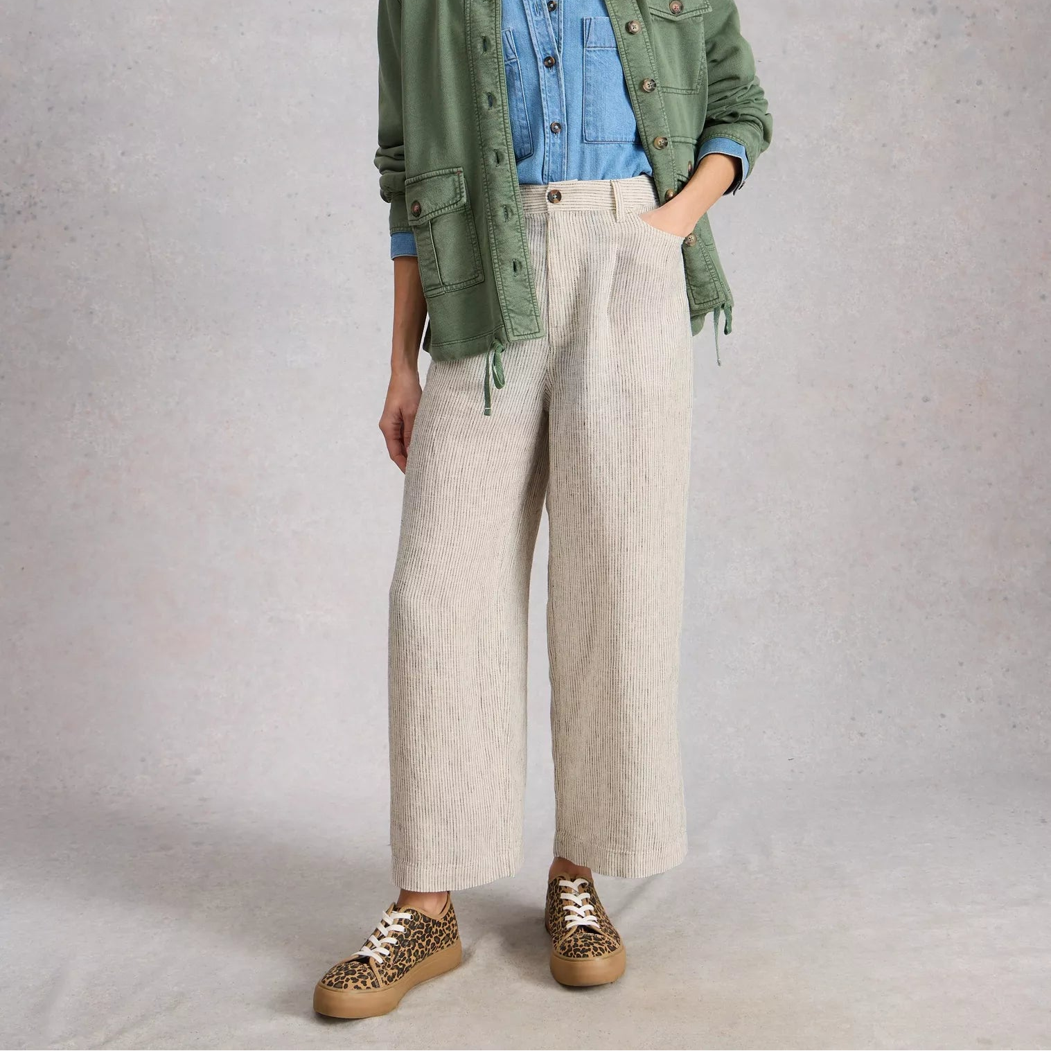 Woman wearing a green jacket, blue shirt, and beige pants against a gray background