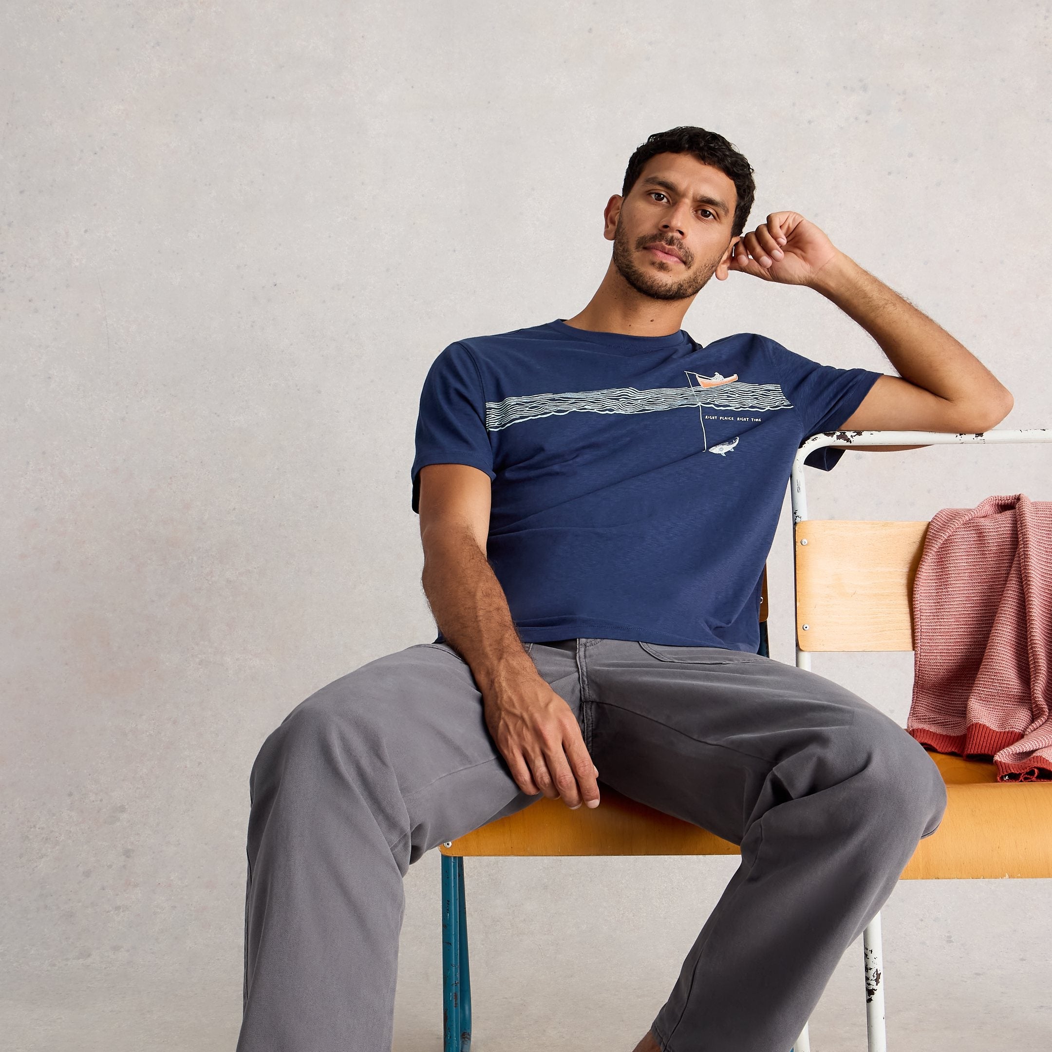 Man sitting on a chair wearing a blue t-shirt and gray pants, with a neutral background.