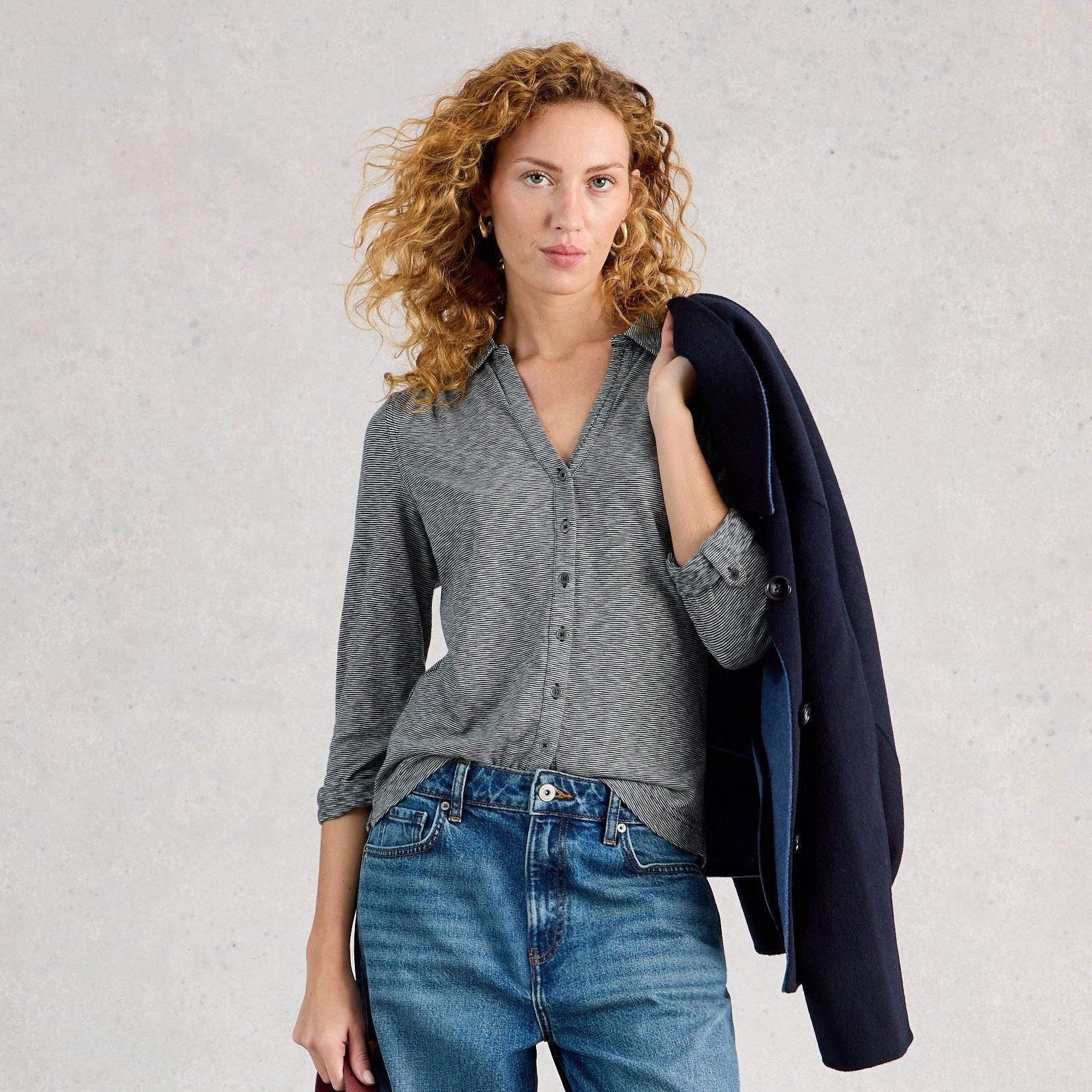 Woman wearing a gray shirt and blue jeans, holding a dark jacket and red bag against a light background