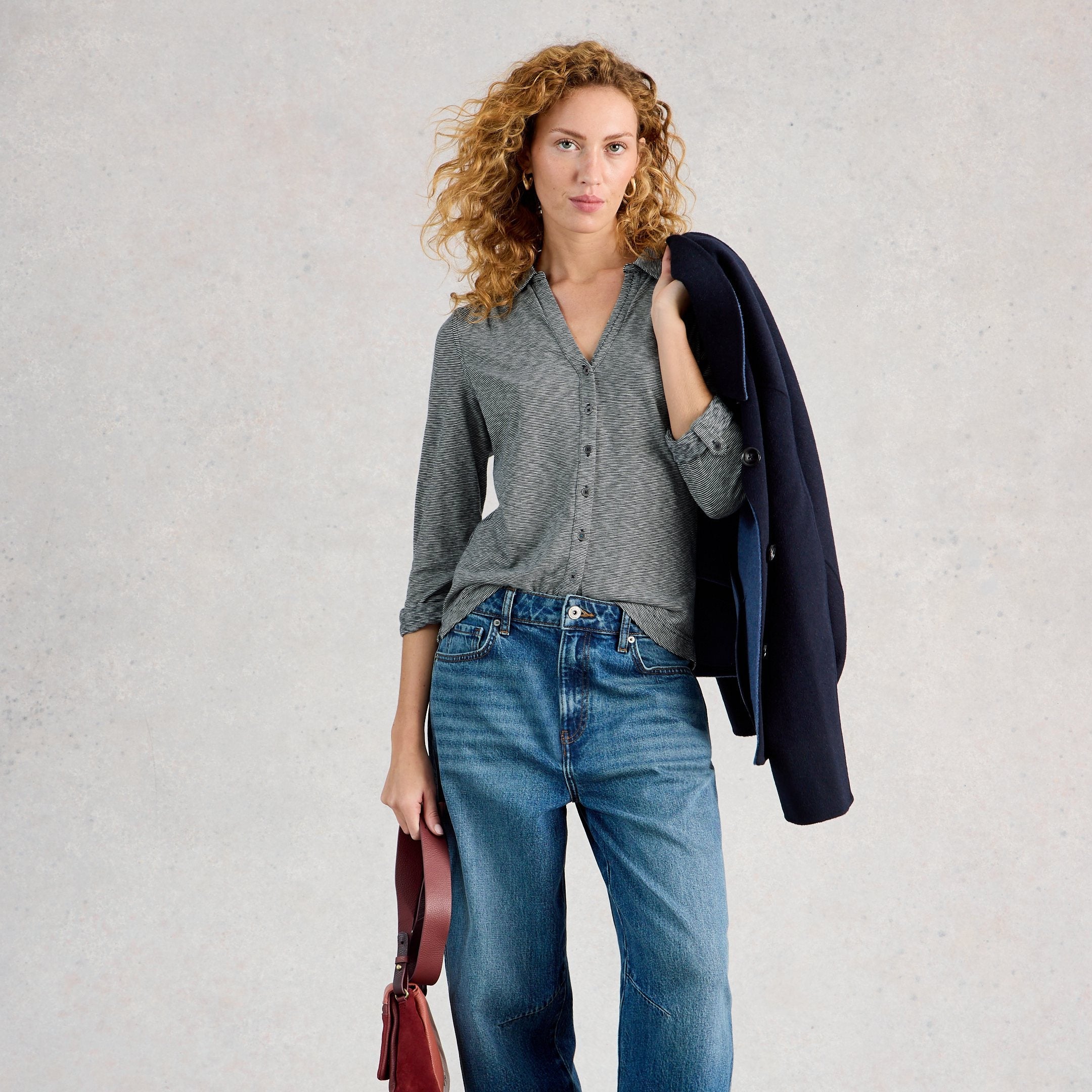 Woman wearing a gray shirt and blue jeans, holding a dark jacket and red bag against a light background