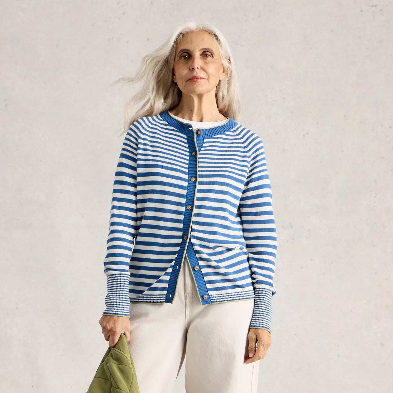 Woman wearing a blue and white striped cardigan with white pants, holding a green jacket against a plain background.