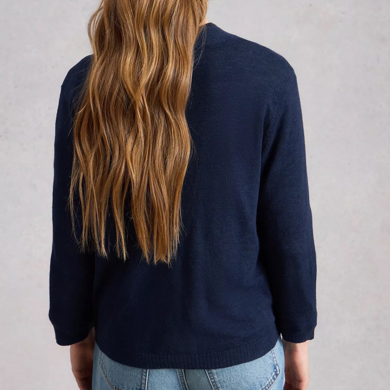 Person wearing a navy sweater and blue jeans with long hair, standing against a plain background