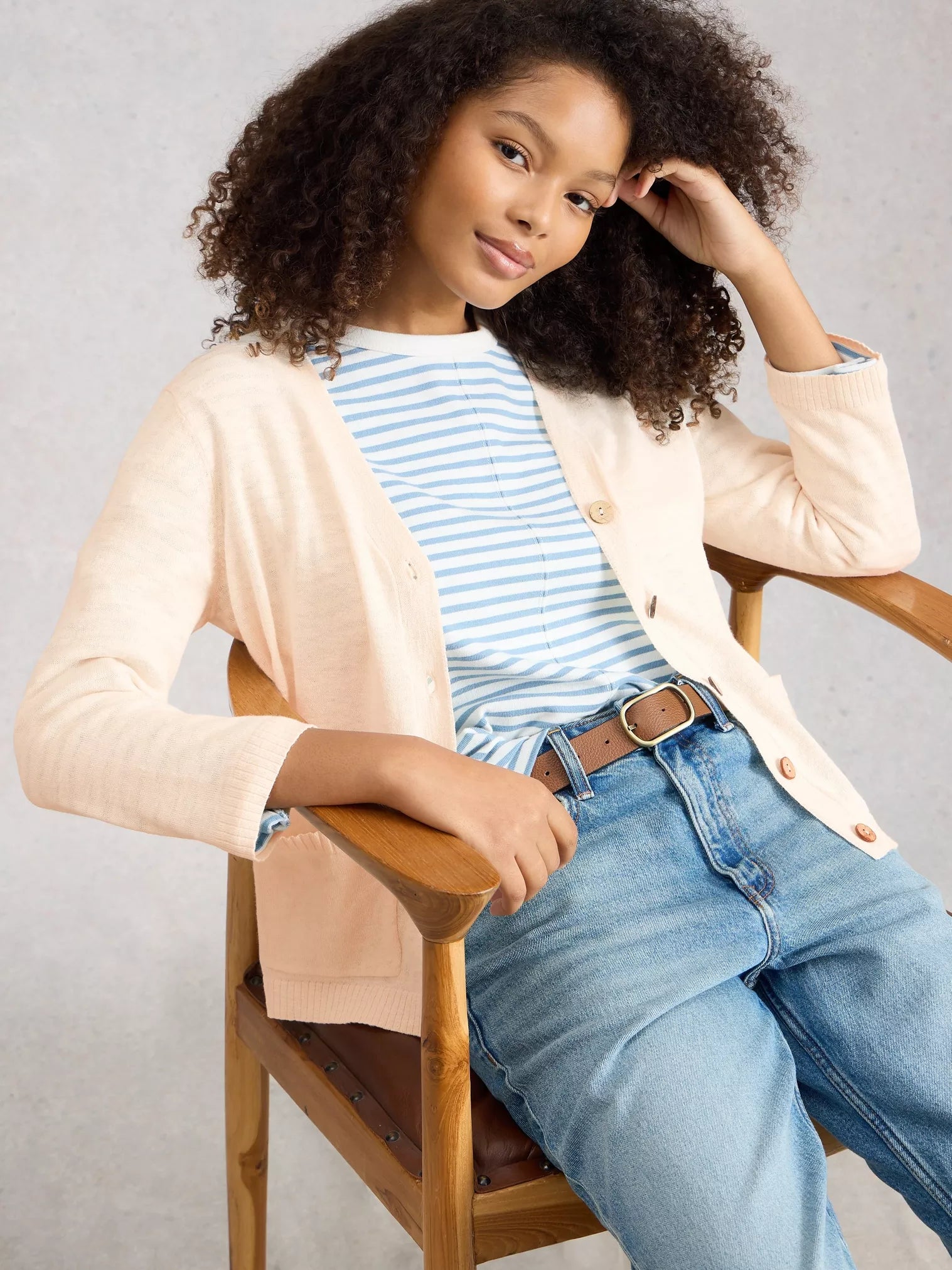 Young girl sitting on a wooden chair wearing a beige cardigan, striped shirt, and jeans.