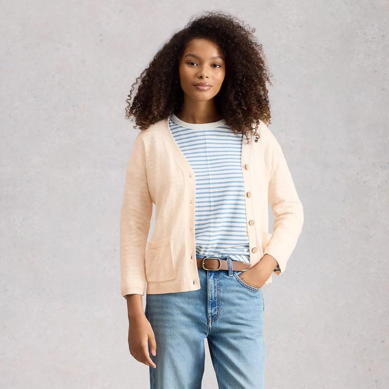 Woman wearing a beige cardigan, striped shirt, and blue jeans on a plain background