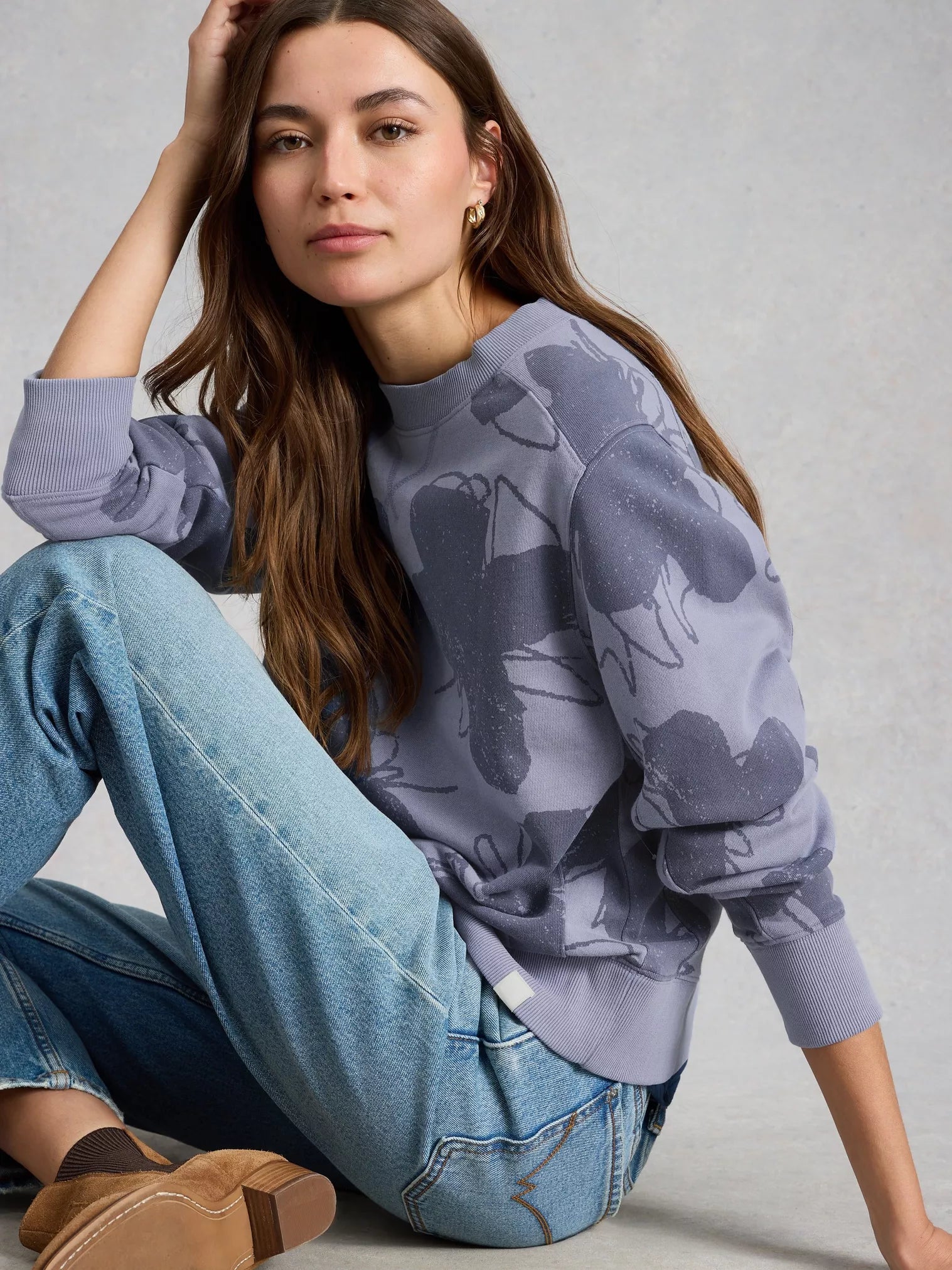 Woman wearing a purple camouflage sweatshirt and blue jeans sitting on the floor against a gray background