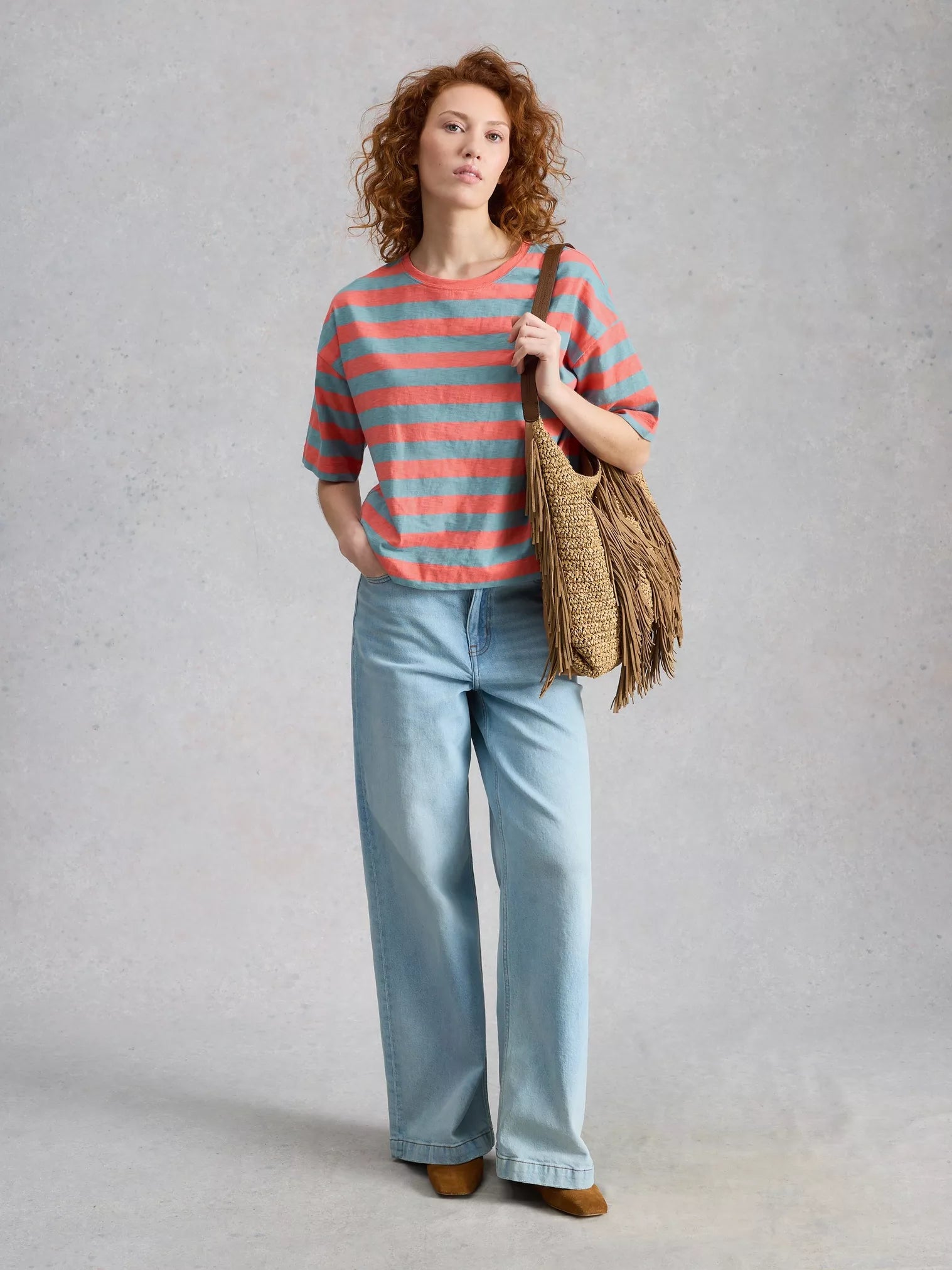 Woman wearing a striped shirt and jeans, holding a woven bag against a plain background