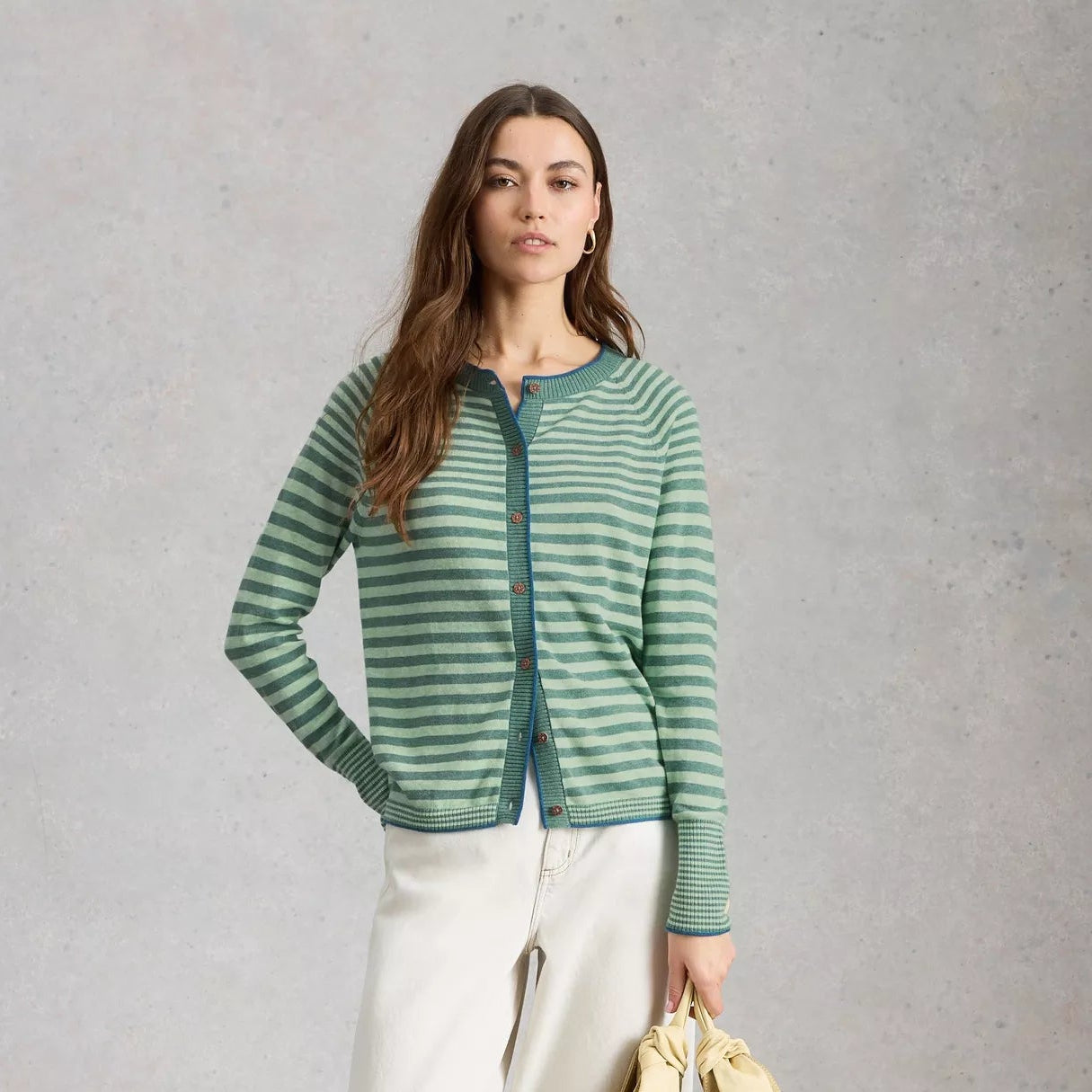 Woman wearing a green striped sweater and white pants, holding a beige bag against a gray background