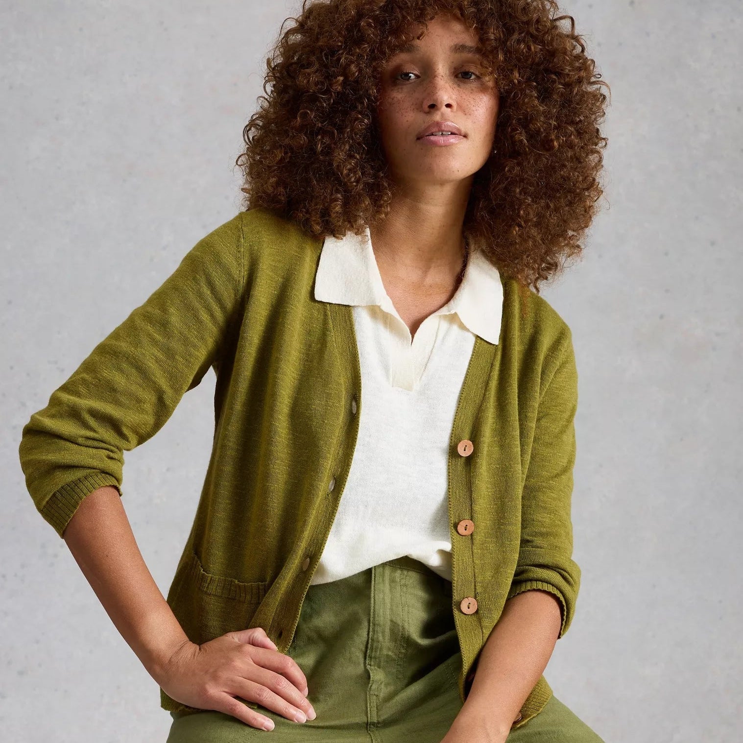 Woman wearing a green cardigan over a white shirt and green pants, sitting on a wooden stool against a gray background.