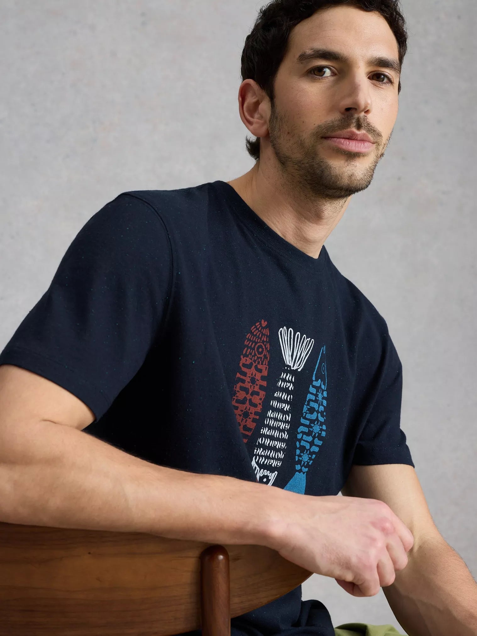 Man wearing a navy blue t-shirt with a graphic design, sitting on a wooden chair against a gray background.