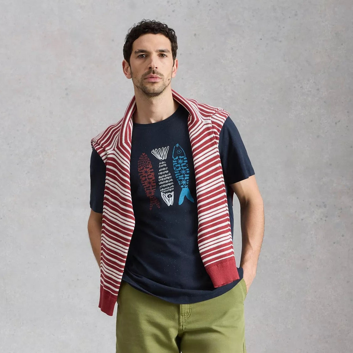 Man wearing a navy t-shirt, green shorts, white sneakers, and a red and white striped vest against a gray background