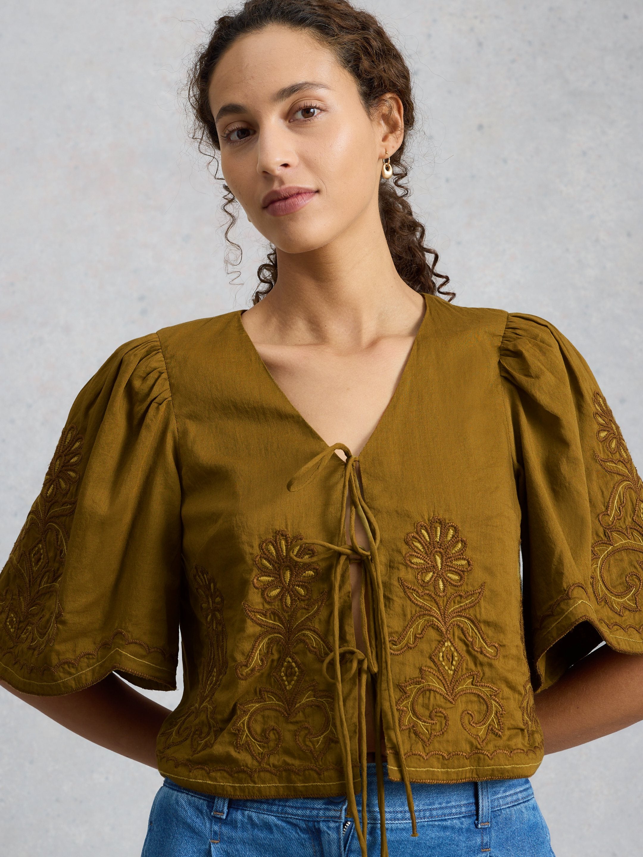 Woman wearing a mustard yellow blouse with floral embroidery on a light gray background