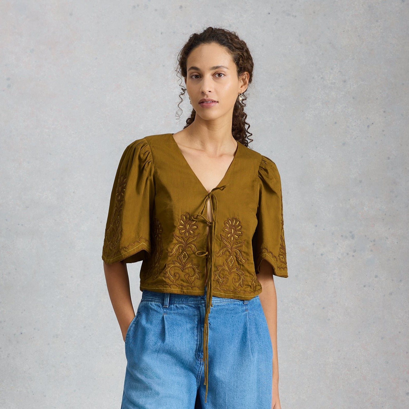 Woman wearing a mustard yellow blouse with floral embroidery and blue denim shorts on a gray background