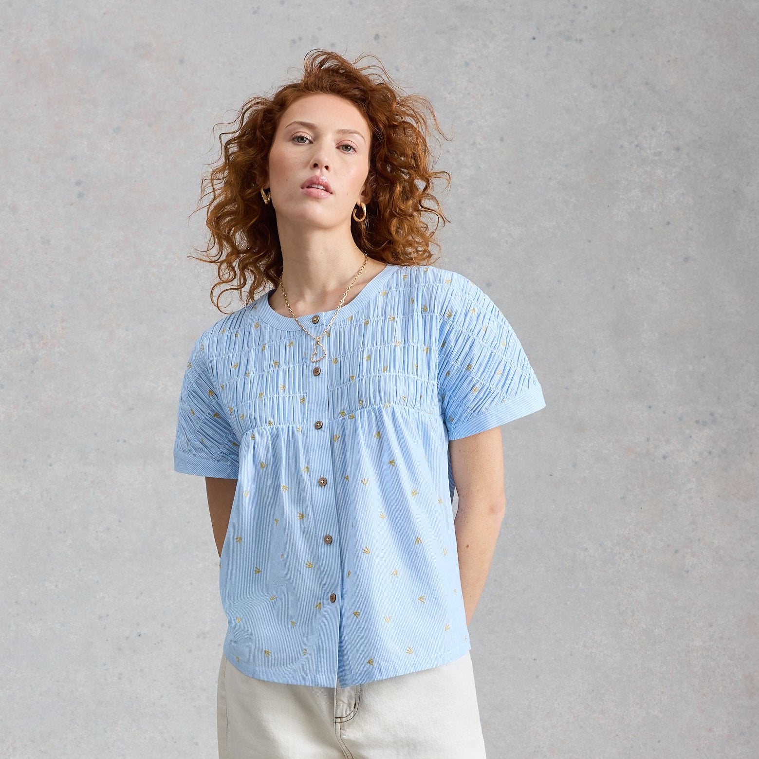 Woman wearing a light blue blouse and beige pants against a gray background