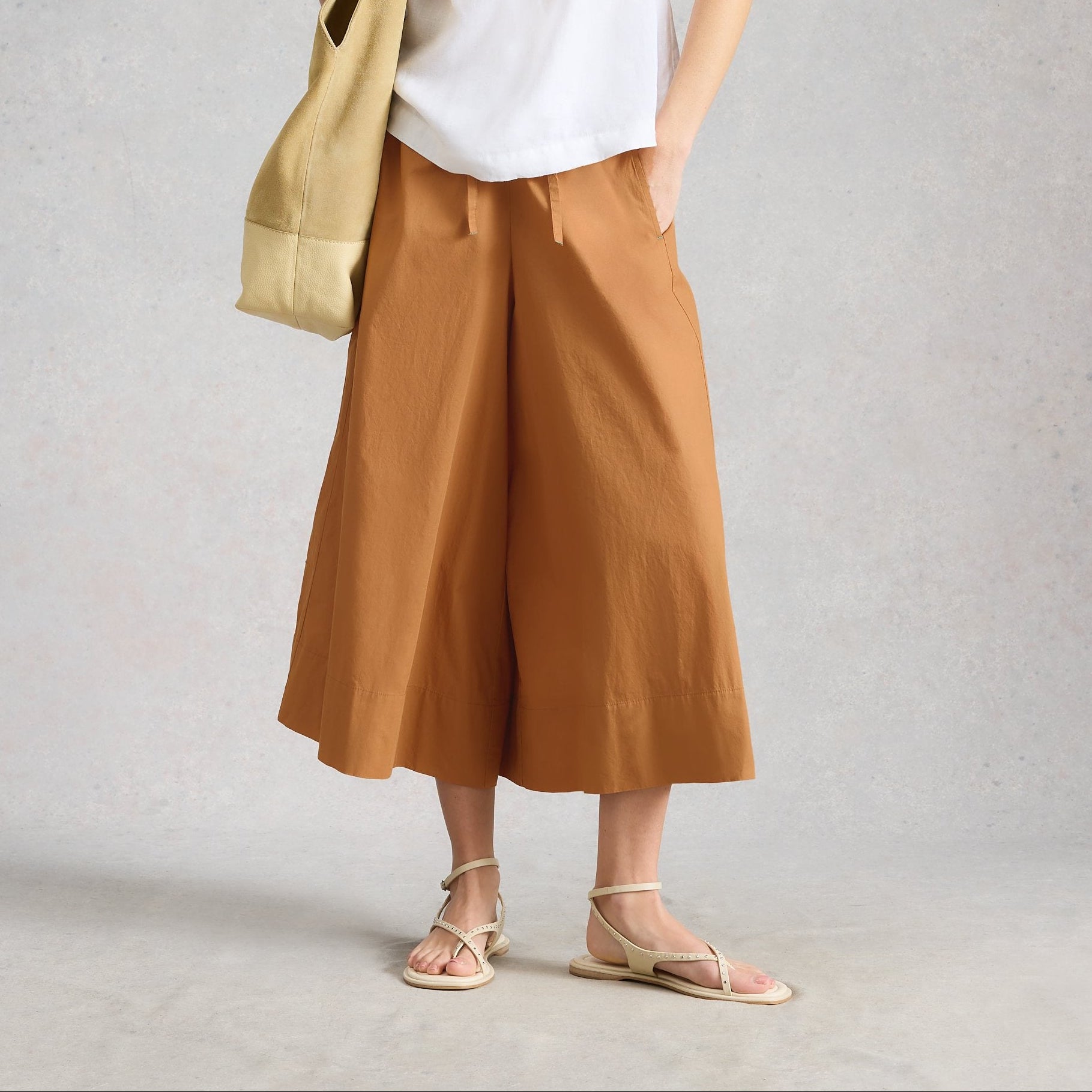 Woman wearing a white sleeveless top and brown wide-leg pants, holding a beige bag against a plain background.