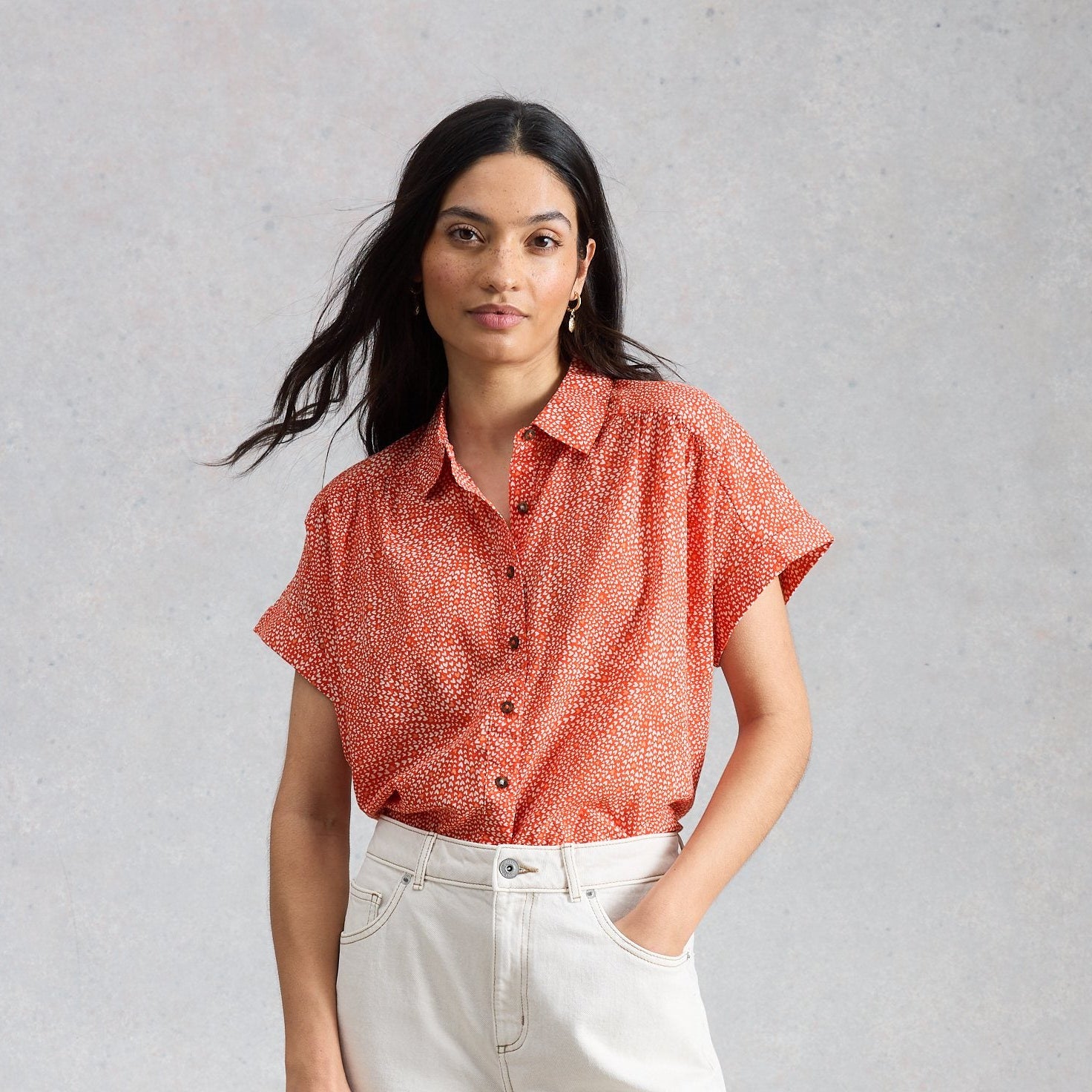 Woman wearing a coral shirt and white pants, holding a woven bag against a gray background