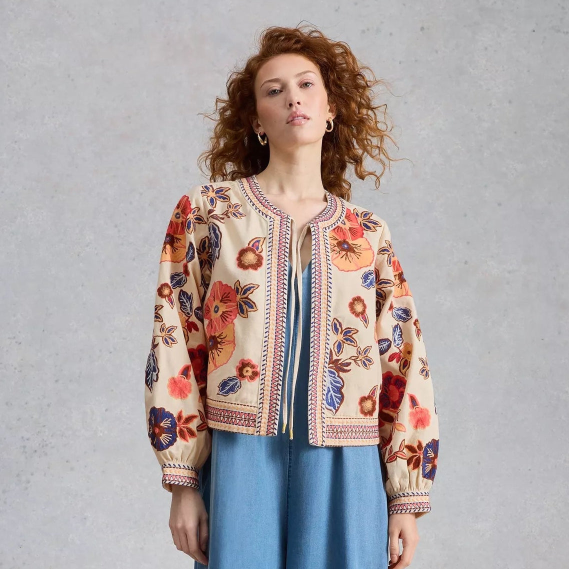 Woman wearing a floral embroidered jacket and blue pants on a gray background