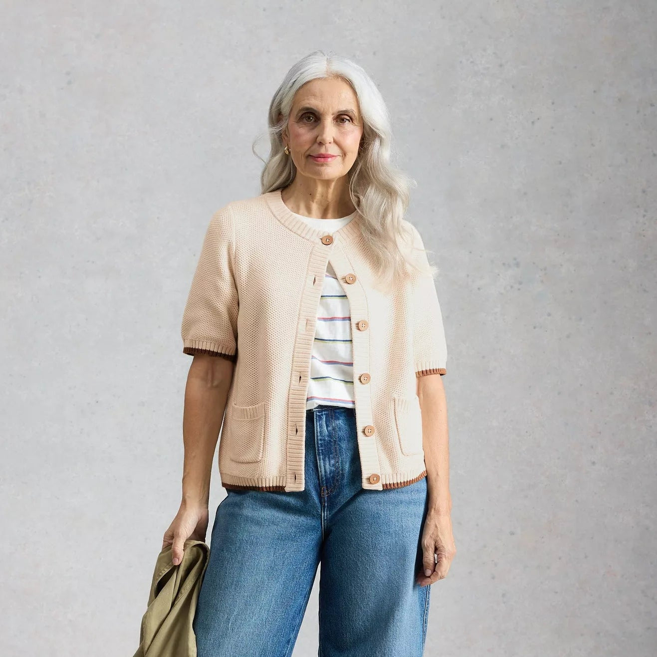 Woman wearing a beige cardigan, blue jeans, and white sneakers on a gray background