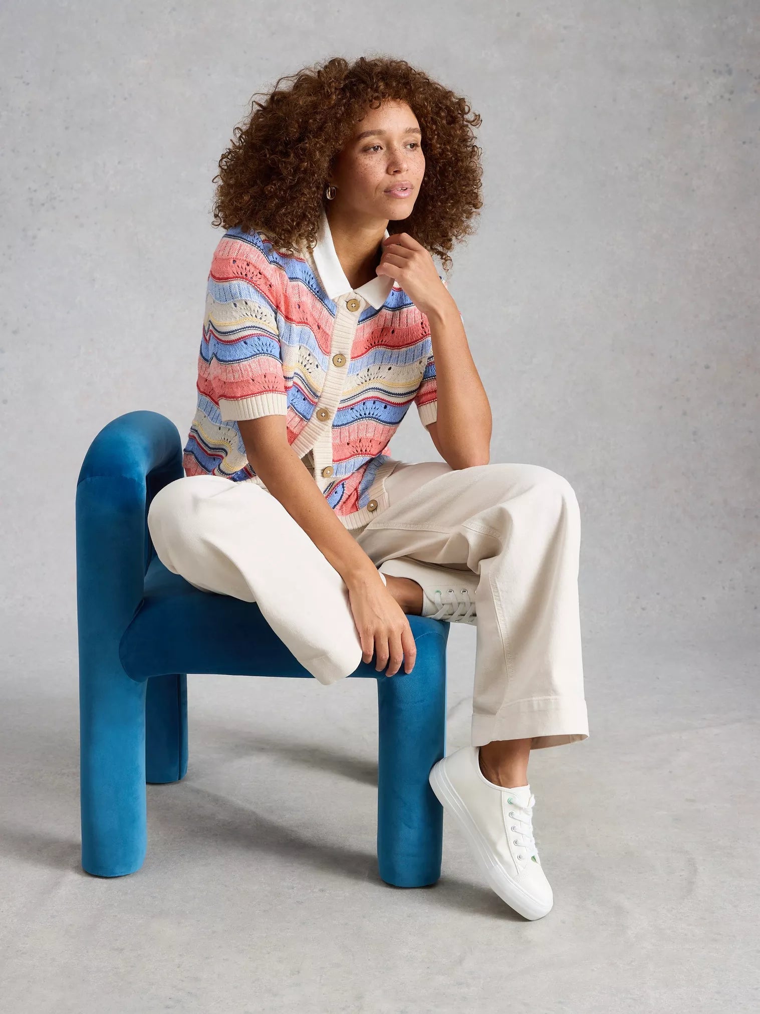 Woman sitting on a blue chair wearing a colorful striped cardigan and white pants against a gray background