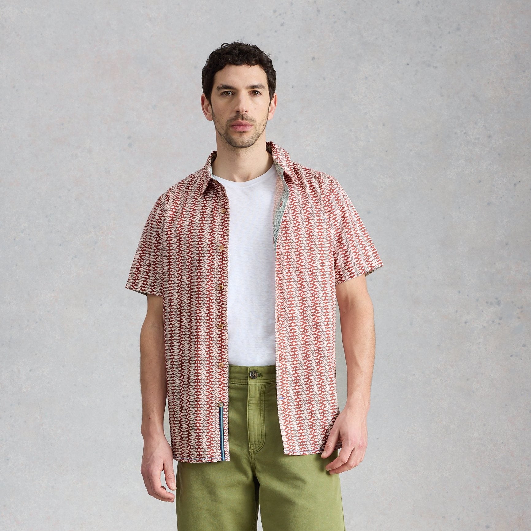 Man wearing a patterned shirt over a white t-shirt with green pants on a gray background