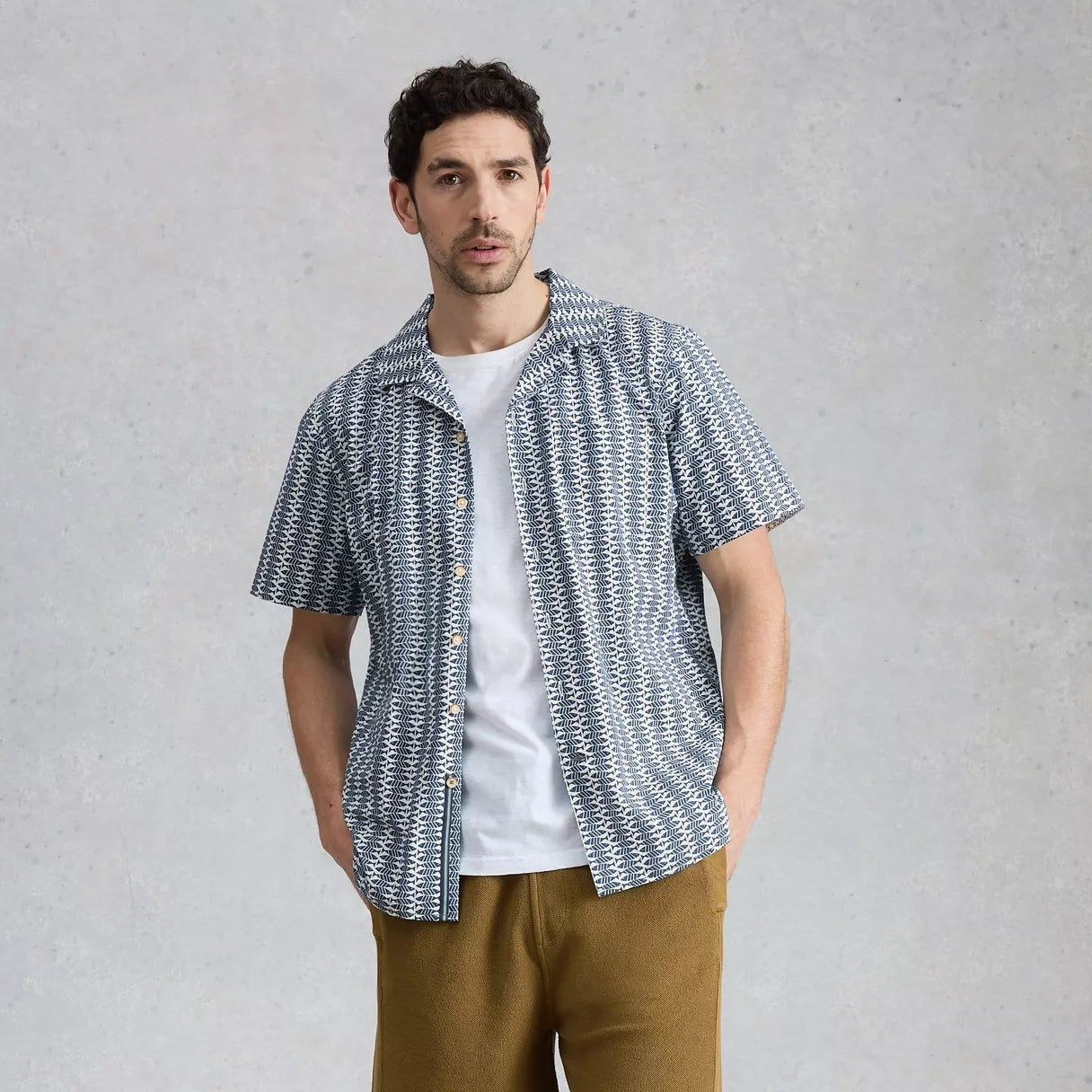 Man wearing a patterned shirt, white t-shirt, and brown shorts against a gray background