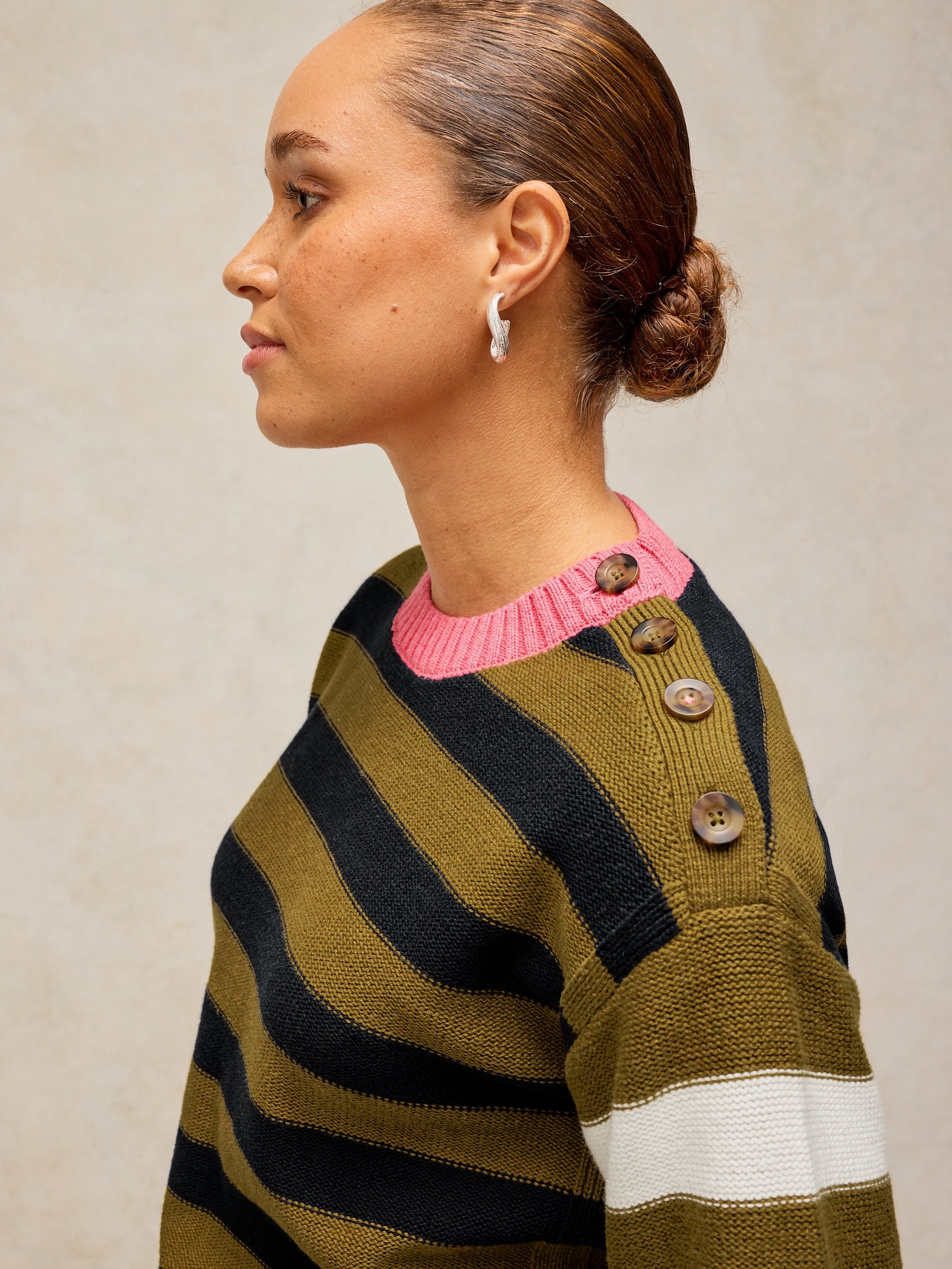 A woman shown in profile wearing a striped knit sweater with olive green, black, and white horizontal stripes, a bright pink crew neck, and brown buttons on the shoulder.