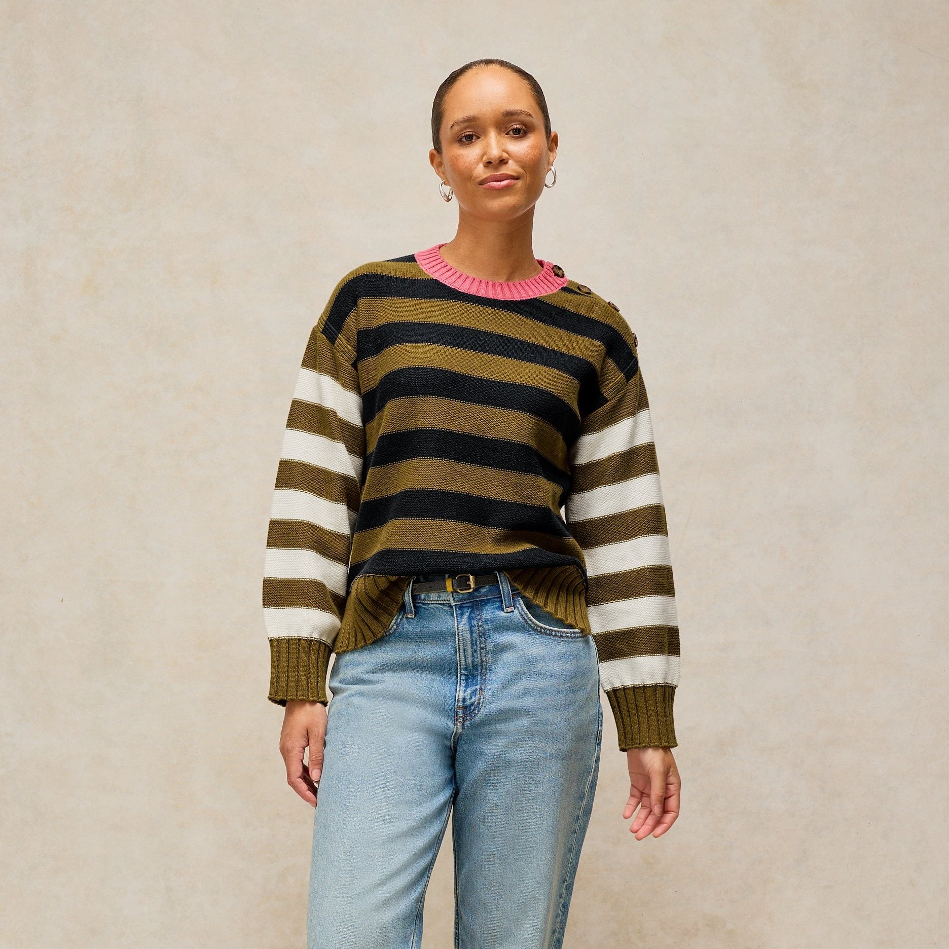 A woman stands against a textured beige studio backdrop wearing a striped knit sweater with olive green, black, and cream horizontal bands, light blue jeans, and brown suede shoes.