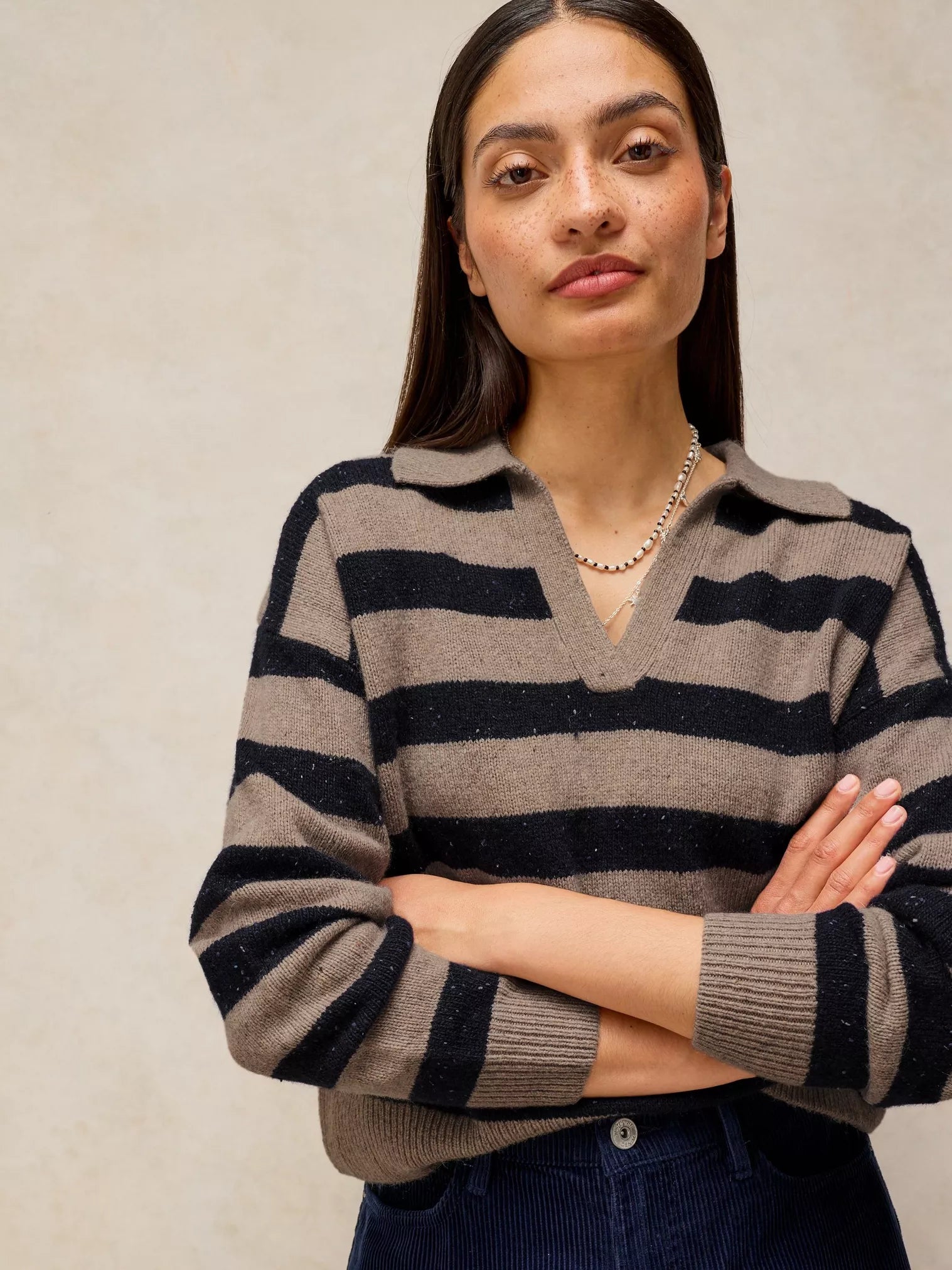 A woman wearing a knit polo-style sweater with horizontal taupe and black stripes, paired with dark corduroy pants, standing against a textured beige wall.