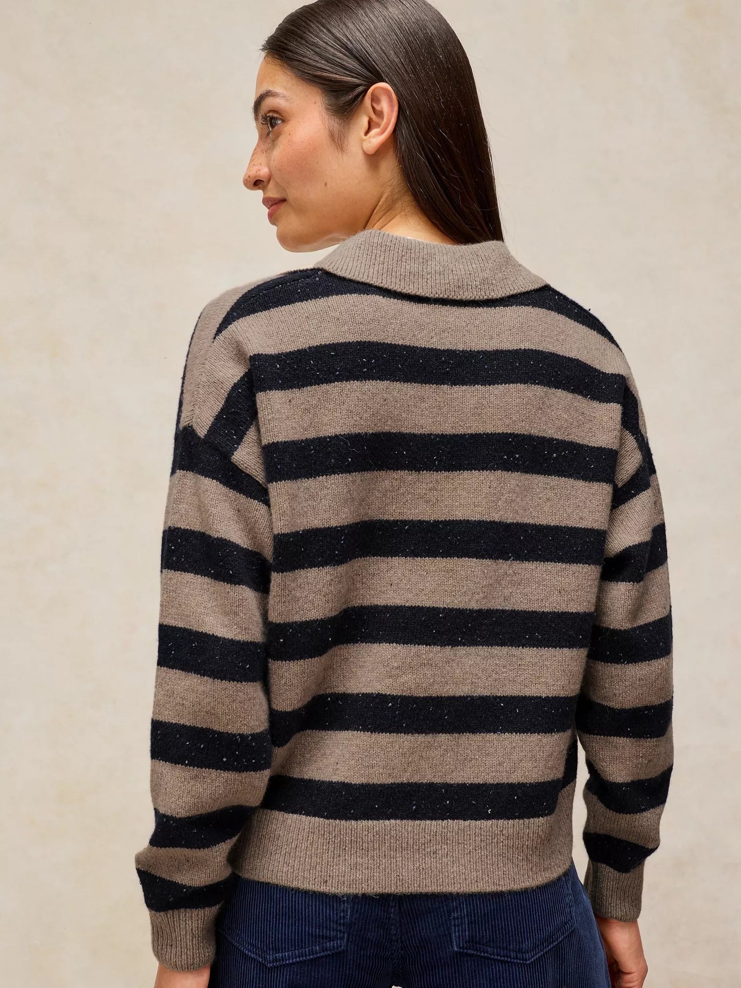 A woman shown from the back wearing a knit sweater with horizontal beige and dark navy stripes, paired with dark blue jeans, against a textured beige background.