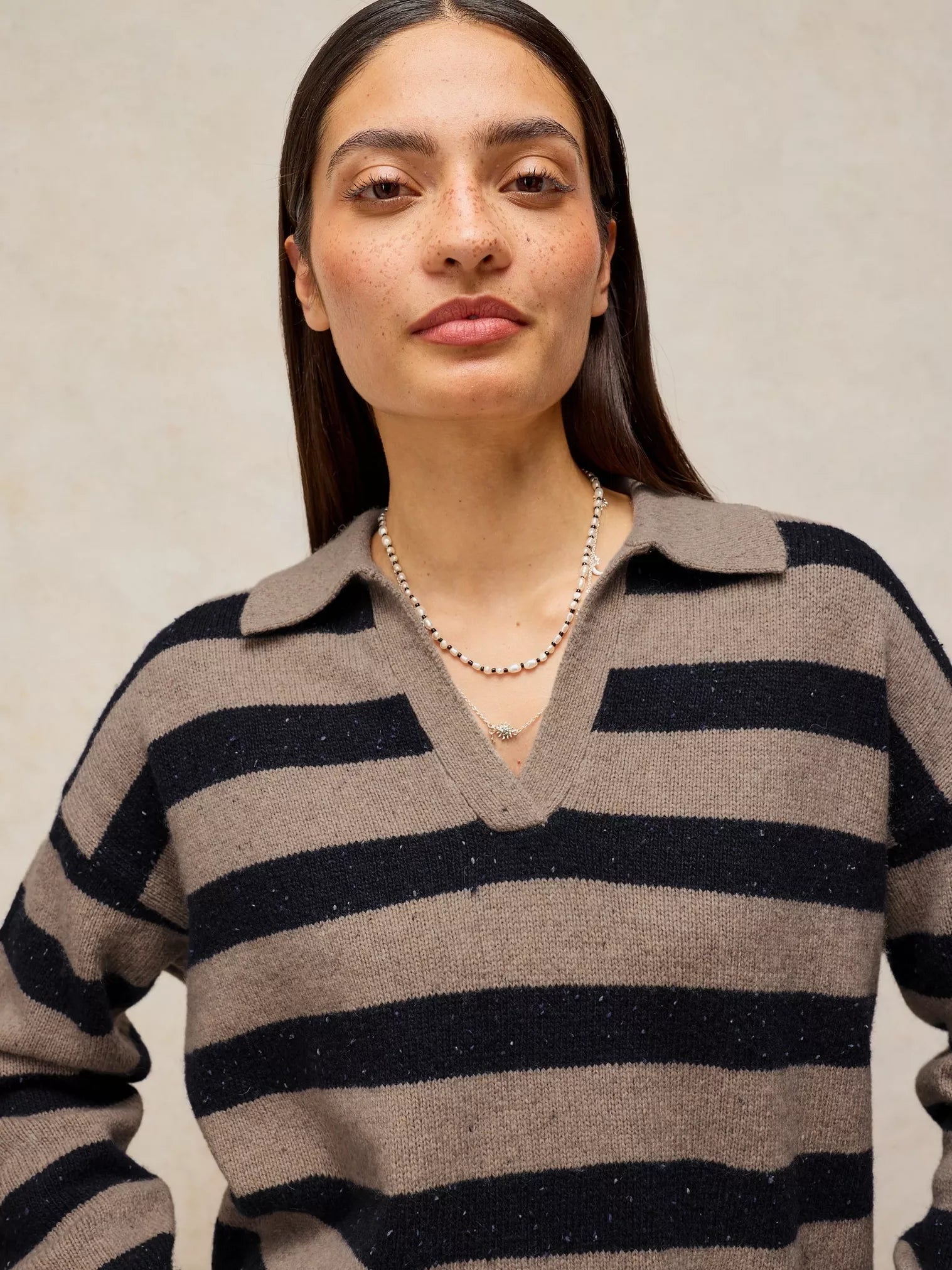 A woman wearing a knit polo-style sweater with horizontal taupe and black stripes, paired with a delicate silver beaded necklace, against a neutral textured beige background.