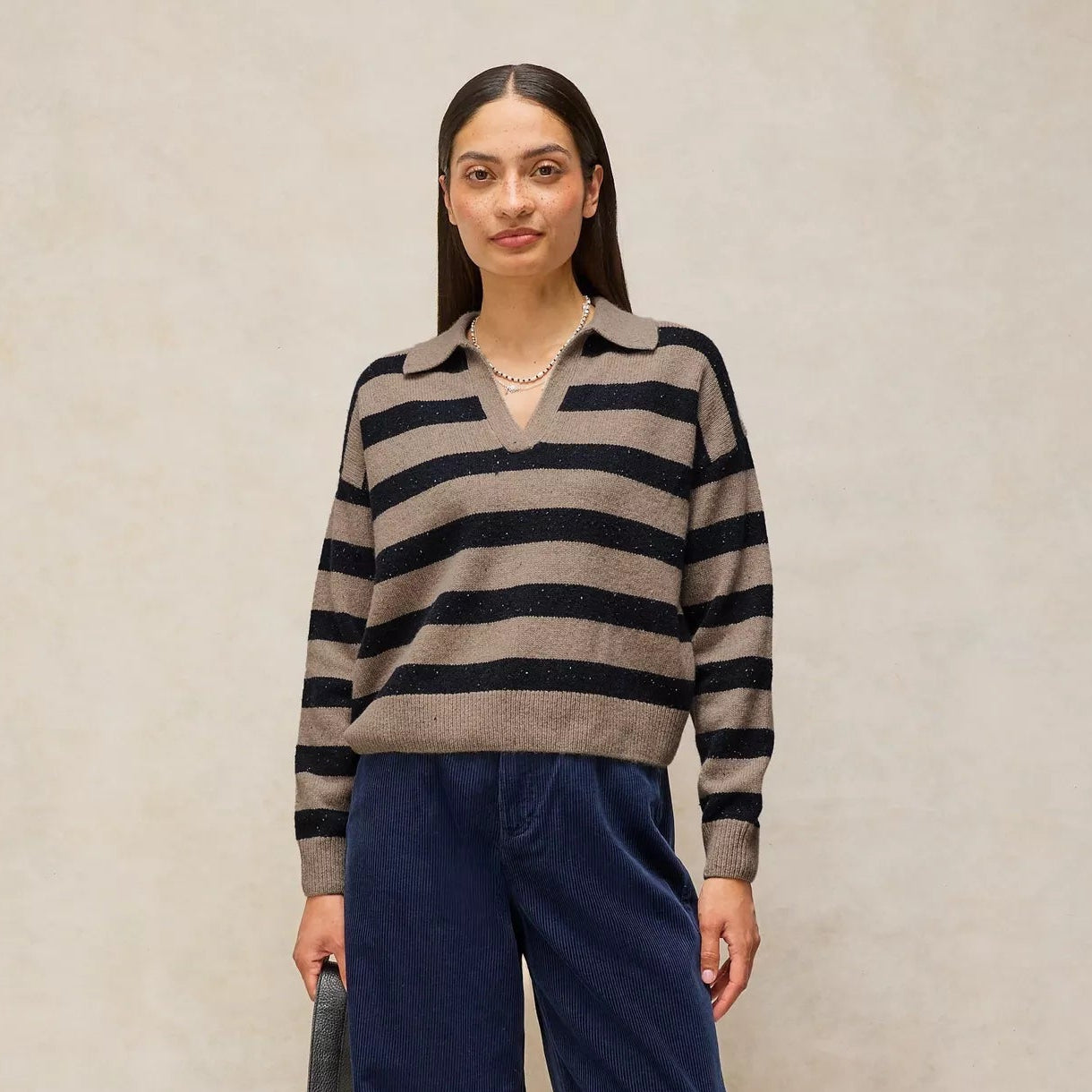 A woman stands against a textured beige studio backdrop wearing a beige and black horizontally striped knit polo sweater, navy wide-leg pants, and black flats, while holding a dark gray shoulder bag.