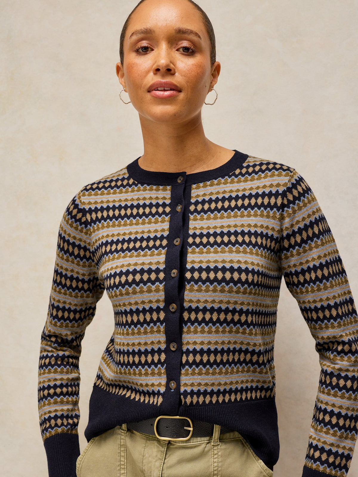 A woman wearing a navy blue button-up cardigan with a beige and navy geometric pattern, paired with khaki pants and a black belt, against a neutral beige background.